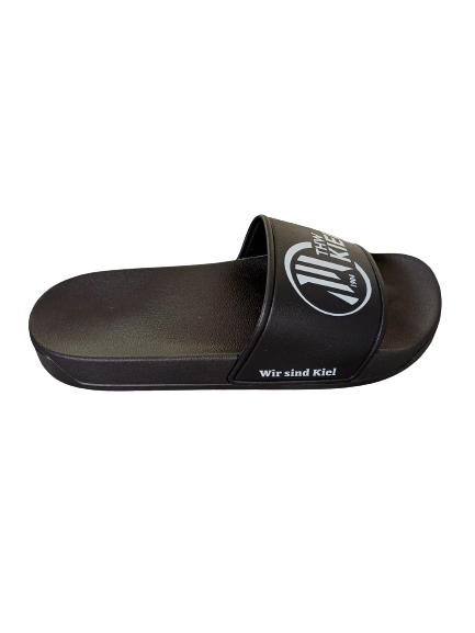 Der Badeschlappen ist eine schwarze Gleitsandale mit weißem Schriftzug "Wir sind Kiel" an der Seite und kreisförmigem Logo auf dem Riemen, ideal für die Sauna, hier auf unifarbenem Hintergrund.