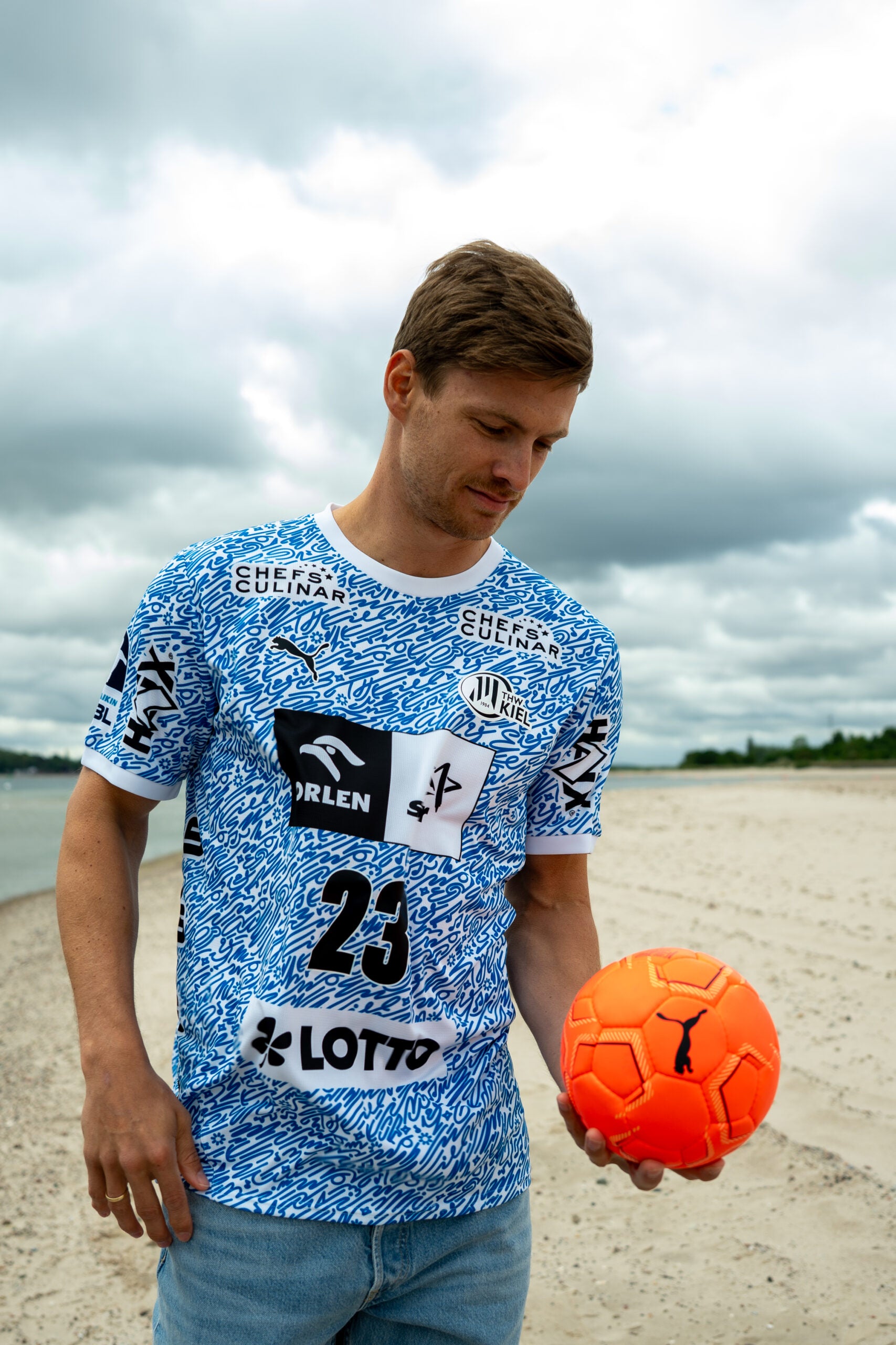 Ein Mann in einem blau-weißen THW Kiel 24-25 Sondertrikot "Kieler Woche" Racing Blue und Jeans steht an einem Sandstrand und schaut nach unten, während er einen orangefarbenen Fußball unter einem bewölkten Himmel hält.