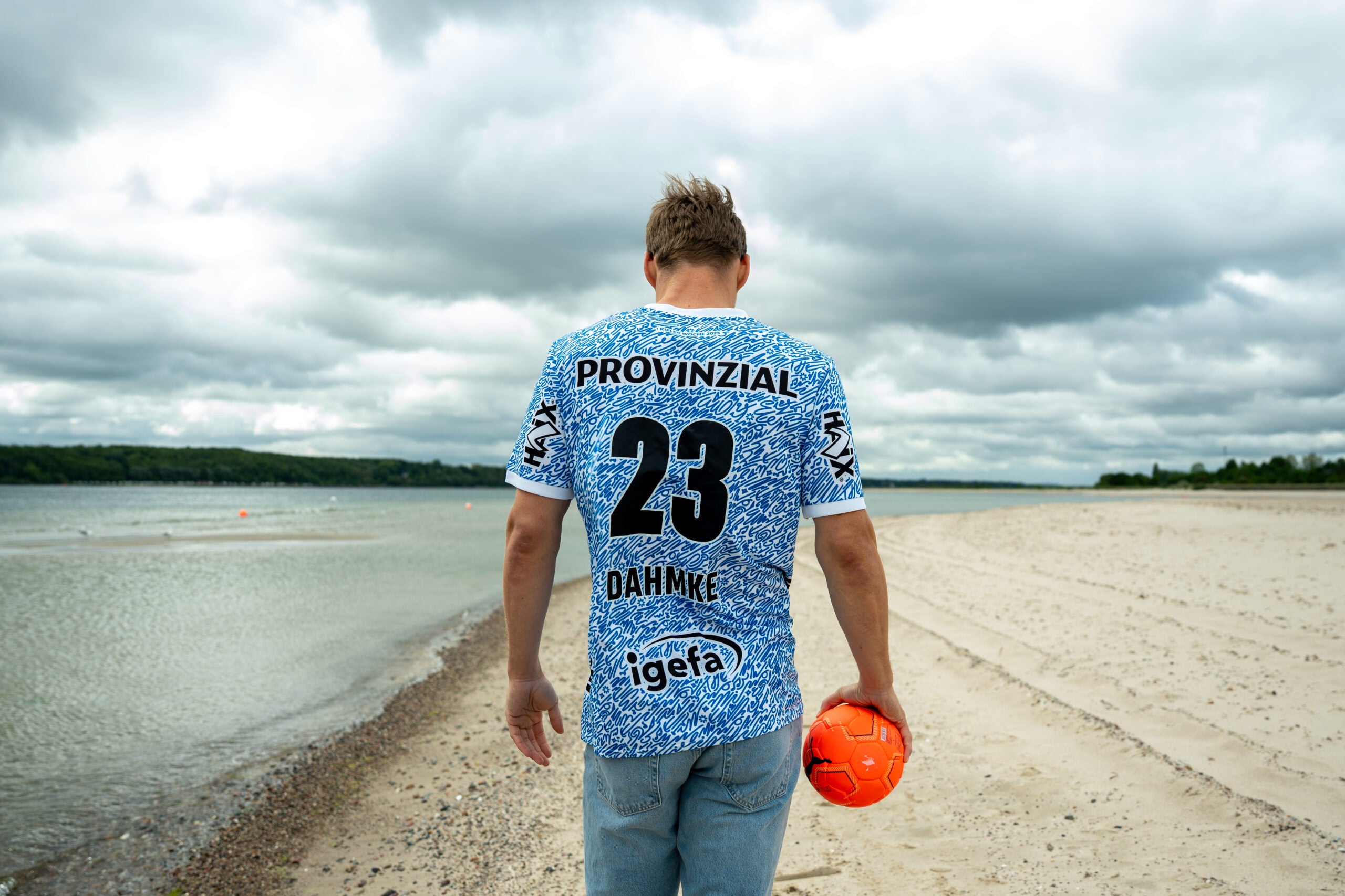 Eine Person in einem THW Kiel 24-25 Sondertrikot "Kieler Woche" Racing Blue mit der Nummer 23 und DAHMKE auf dem Rücken läuft über einen Sandstrand, in der Hand einen orangefarbenen Ball, mit Wasser und bewölktem Himmel im Hintergrund.