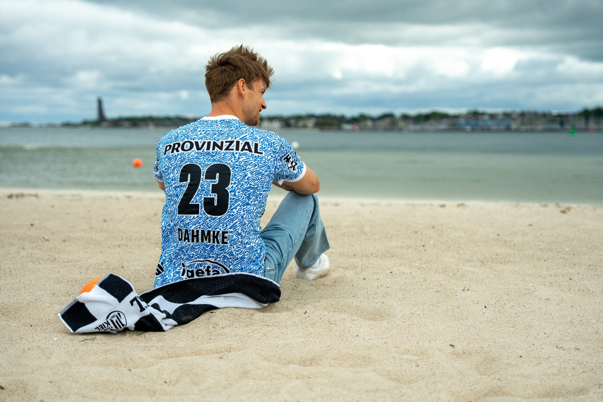Ein Mann im blau-weißen THW Kiel 24-25 Sondertrikot Jr "Kieler Woche" Racing Blue mit der 23 Dahmke auf dem Rücken sitzt bei bewölktem Himmel am Sandstrand, ein schwarz-weißes Handtuch neben sich - vielleicht in Erwartung der Kieler Woche 2025.