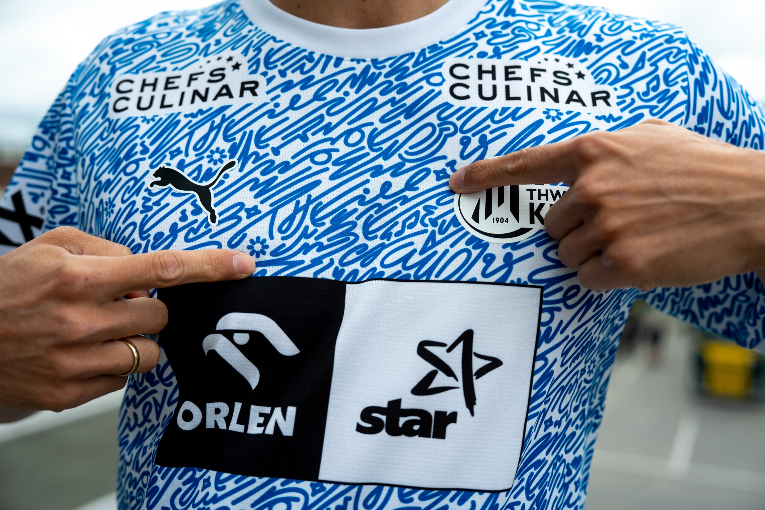 Eine Person, die das THW Kiel 24-25 Spezialtrikot Jr "Kieler Woche" Racing Blue trägt, mit blau-weißem Muster, mehreren Sponsorenlogos und einem runden Brustabzeichen, möglicherweise für die Kieler Woche 2025 entworfen.