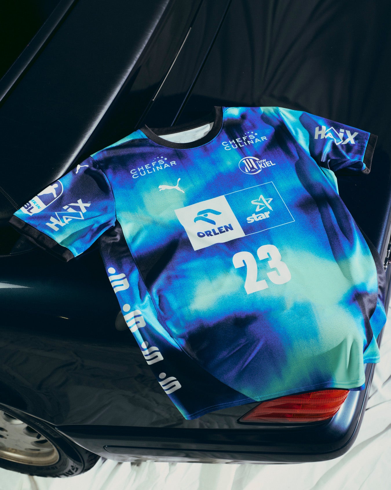 Ein navyfarbenes THW Kiel 25-26 Away Jersey Jr by PUMA mit Sponsorenlogos und der Nummer 23 ist über das Heck eines schwarzen Autos drapiert.