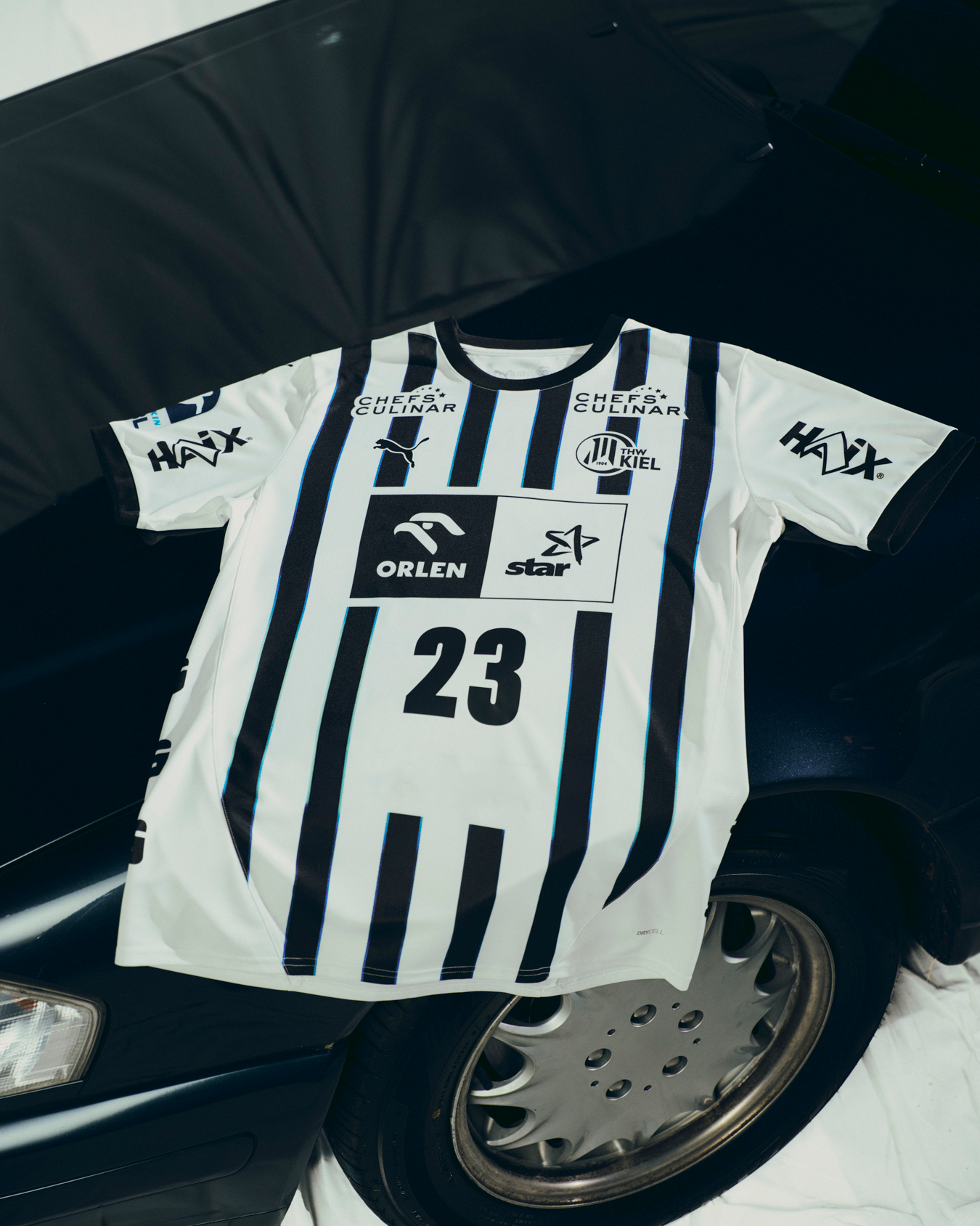 Das THW Kiel 25-26 Heimtrikot Jr PUMA White mit schwarzen Streifen, der Nummer 23 und Sponsorenlogos ist auf der Motorhaube eines dunklen Autos abgebildet und verdeckt teilweise die Windschutzscheibe und den Vorderreifen.