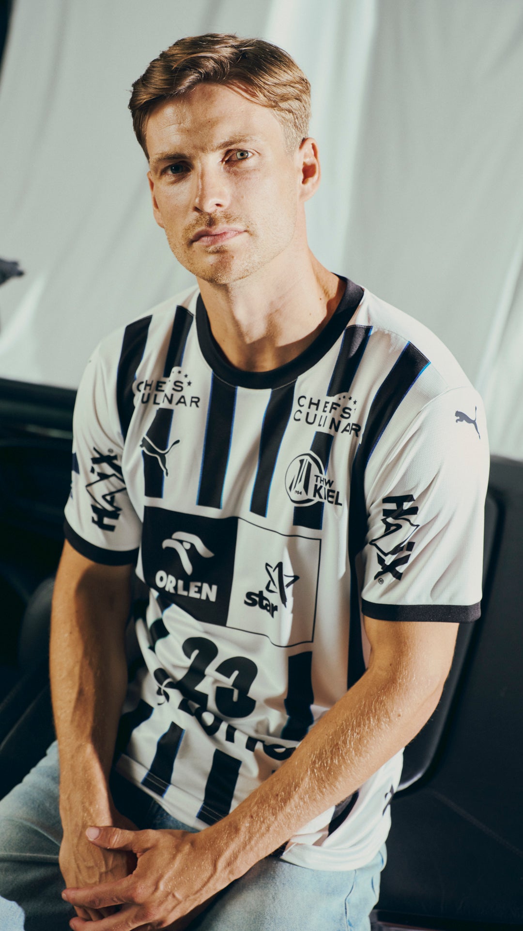 Ein Mann mit kurzen hellbraunen Haaren sitzt vor einem schlichten Hintergrund, blickt in die Kamera und trägt das THW Kiel 25-26 Heimtrikot Jr PUMA White mit schwarzen Streifen und Sponsorenlogos sowie eine hellblaue Jeans.