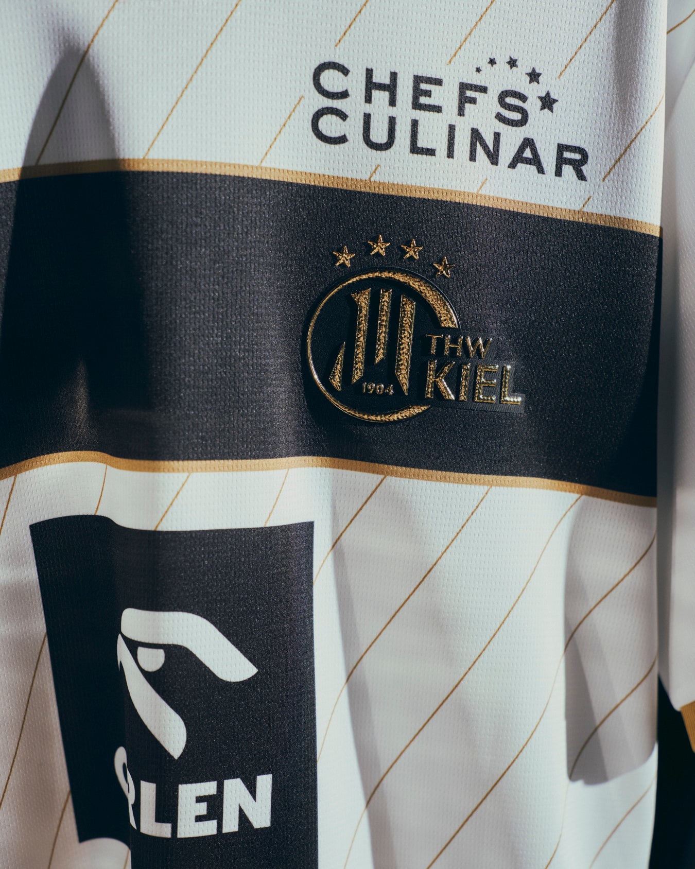 Eine Nahaufnahme des THW Kiel 25-26 Euro Home Trikots Jr von PUMA in Weiß mit dem Mannschaftswappen, den Sponsorenlogos von CHEFS CULINAR und GLEN sowie dezenten diagonalen Linien in Gold auf weißem und schwarzem Hintergrund.