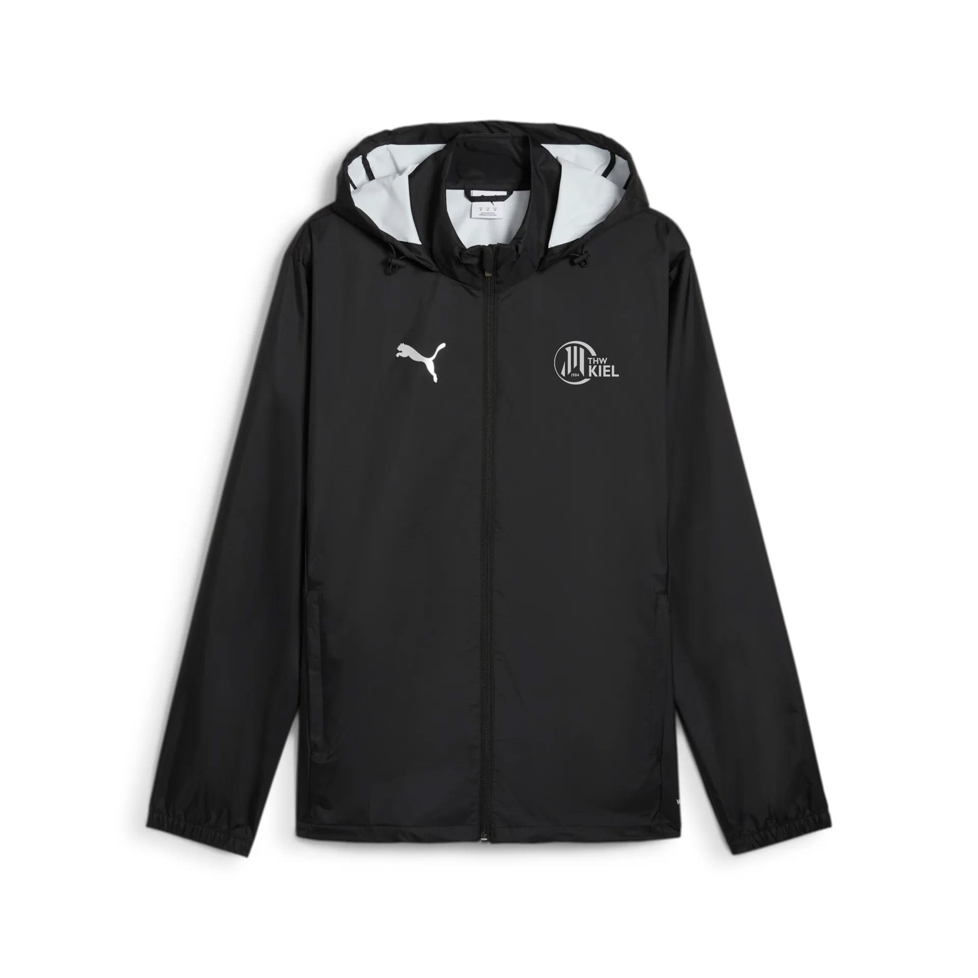 Die teamADDITIONS Training All Weather Jacket in PUMA Schwarz zeigt ein weißes Puma-Logo auf der rechten Brust und ein kreisförmiges Vereinslogo von Holstein Kiel auf der linken Seite, das auf einem schlichten weißen Hintergrund abgebildet ist.