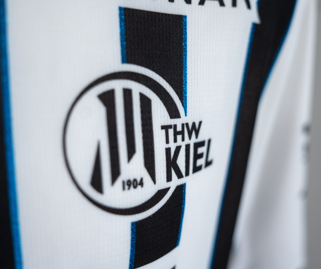 Nahaufnahme des THW Kiel 25-26 Heimtrikots in PUMA-Weiß. Es zeigt das kultige schwarz-weiße THW Kiel-Logo mit "THW KIEL" und "1904" auf einem runden Emblem über stylischen schwarzen und blauen Längsstreifen.