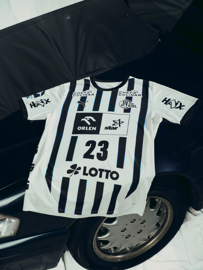 Das THW Kiel 25-26 Heimtrikot Jr PUMA White mit schwarzen Streifen, der Nummer 23 und den Sponsorenlogos von ORLEN und LOTTO liegt flach auf der Motorhaube eines dunklen Autos mit einem sichtbaren Rad.