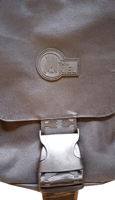 Eine Nahaufnahme der Cross Body Bag BUCKLE schwarz mit Kunststoffschnalle, Reißverschluss und erhabenem, kreisförmigem THW Kiel Logo auf der Klappe.