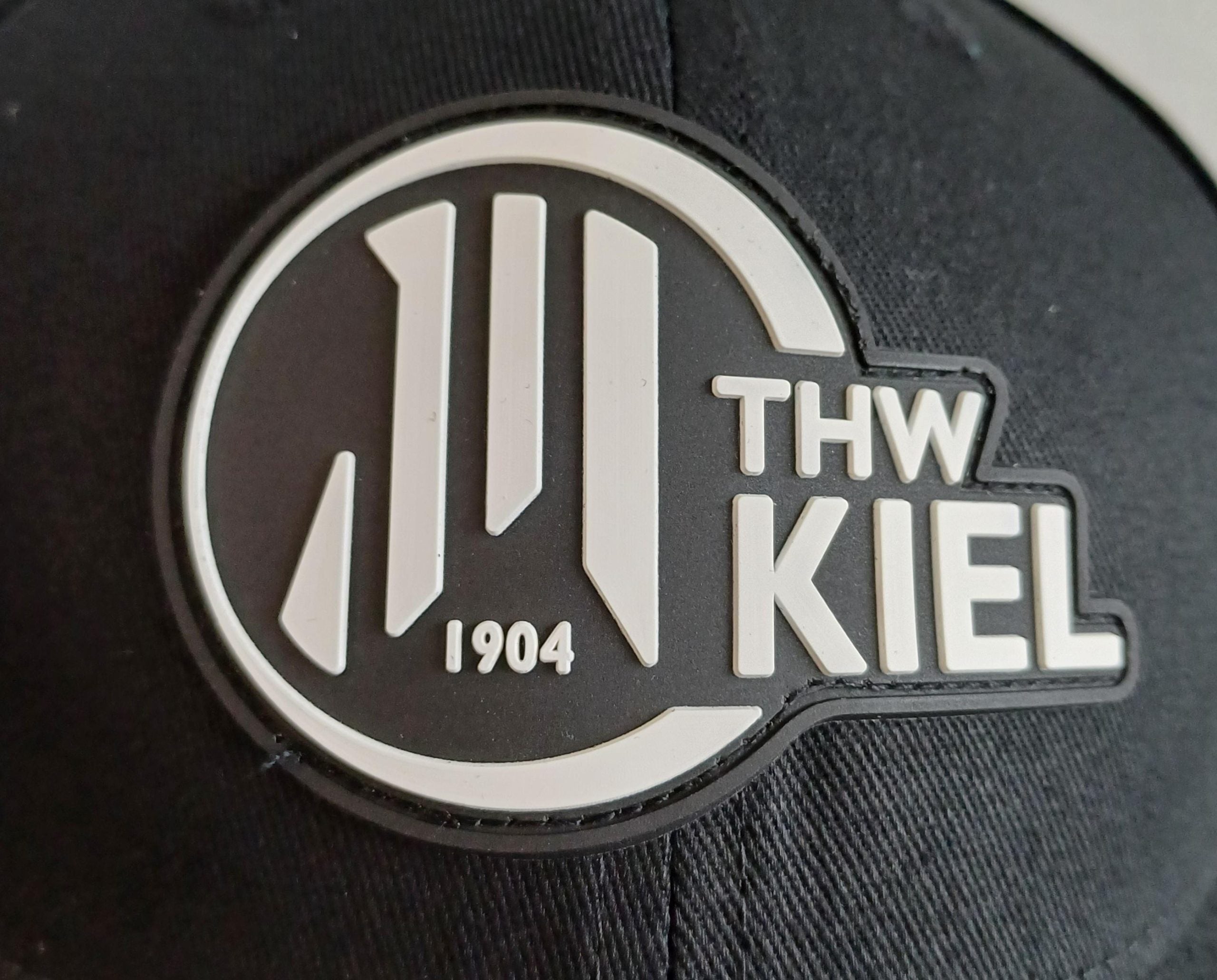 Der Cap Rubber Patch schwarz zeigt die Nahaufnahme einer schwarzen Kappe mit dem THW Kiel-Logo in Weiß als 3D-Gummiaufnäher, auf dem "THW KIEL" und "1904" neben vertikalen Linien in einem kreisförmigen Design zu sehen sind.