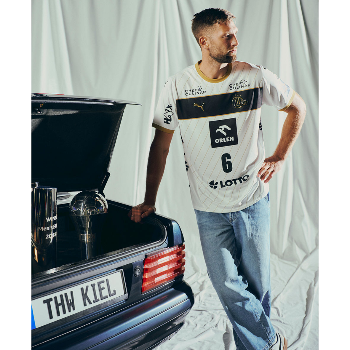 Ein Mann, der das THW Kiel 25-26 Euro Home Jersey PUMA White und eine blaue Jeans trägt, lehnt an einem offenen Kofferraum mit zwei Trophäen, einem weißen Stoffhintergrund hinter ihm und einem Nummernschild mit der Aufschrift THW KIEL.