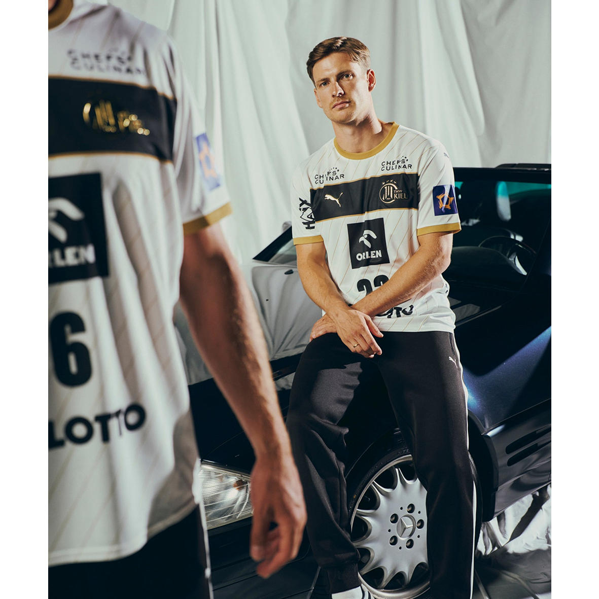 Ein Mann, der das THW Kiel 25-26 Euro Home Jersey PUMA White trägt, sitzt auf einem schwarzen Auto und schaut in die Kamera. Eine weitere Person in einem ähnlichen Trikot steht im Vordergrund, teilweise außerhalb des Bildausschnitts.