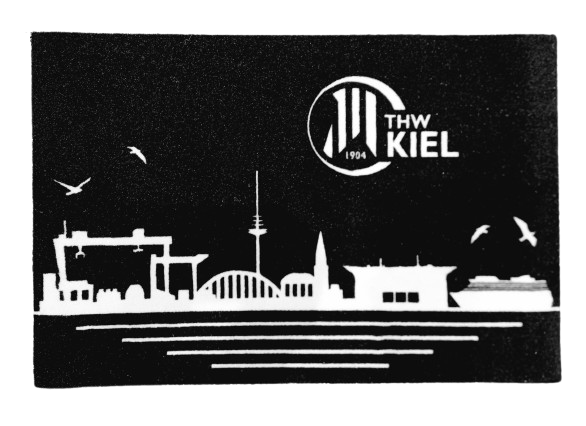 Die Fußmatte Skyline schwarz zeigt eine weiße Silhouette der Kieler Skyline - Gebäude, Brücke, Schiff und Möwen - sowie das THW Kiel Logo und 1904 auf schwarzem Hintergrund. Eine stilvolle, rutschfeste Fußmatte für Fans!.