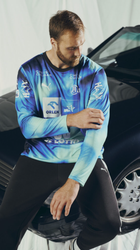 Ein Mann, der ein THW Kiel 25-26 GK Trikot Home PUMA Navy mit Sponsorenlogos trägt, sitzt auf der Motorhaube eines schwarzen Autos, schaut nach unten und richtet seinen Ärmel.