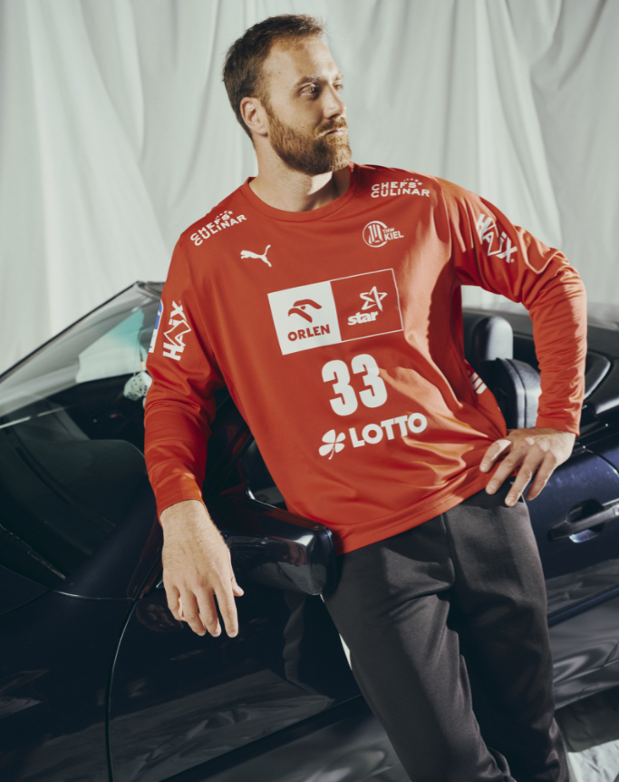Ein bärtiger Mann in einem roten THW Kiel 25-26 GK Trikot von PUMA, mit "33" und Logos, lehnt an einem dunklen Auto. Weißer drapierter Stoff im Hintergrund unterstreicht diesen Look aus 100% Polyester der Saison 2025/26.