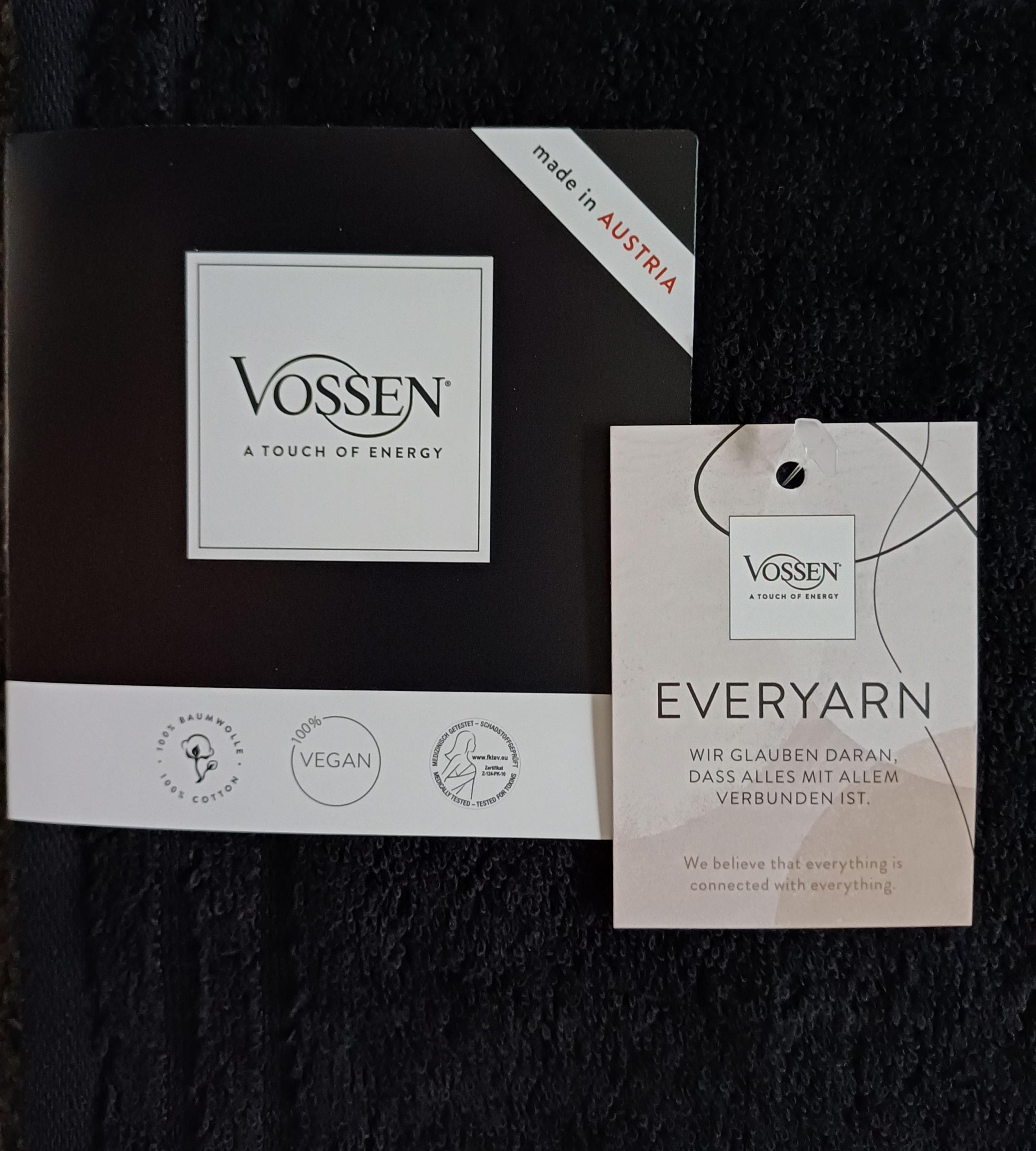 Das Handtuch 50 x 100cm schwarz trägt den Slogan "A Touch of Energy", vegane und nachhaltige Baumwollsymbole, ein EVERYARN-Tag und "Wir glauben, dass alles mit allem verbunden ist" in Englisch und Deutsch.