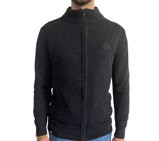 Eine Person trägt den Strickcardigan, einen schwarzen Pullover aus Merinowolle mit Reißverschluss, hohem Kragen und dezentem Logo auf der linken Brustseite, kombiniert mit einer hellblauen Jeans. Nur der Oberkörper ist sichtbar.