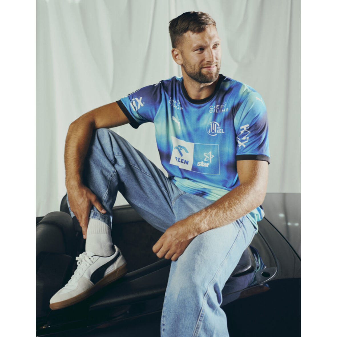 Ein Mann mit kurzen braunen Haaren und Bart, bekleidet mit dem THW Kiel 25-26 Auswärtstrikot PUMA Navy, hellen Jeans, weißen Socken und weißen Turnschuhen sitzt lässig mit einem angehobenen Knie auf einem schwarzen Auto, schaut zur Seite und lächelt.