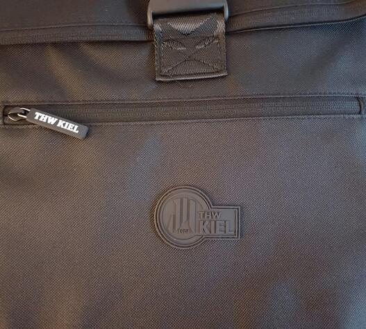 Die Nahaufnahme des Rucksack BUCKLE schwarz zeigt einen sichtbaren Reißverschluss mit THW KIEL-Anhänger, ein geprägtes THW KIEL-Logo auf der Oberfläche und ein praktisches Laptop-Fach im Inneren.