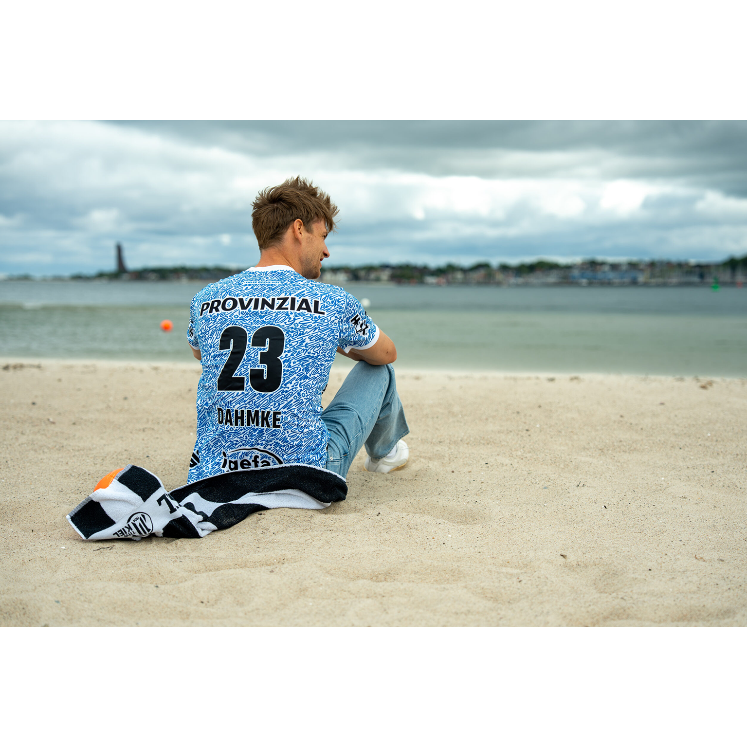 Ein Mann sitzt während der Kieler Woche 2025 an einem Sandstrand und trägt das THW Kiel 24-25 Sondertrikot Jr "Kieler Woche" Racing Blue mit PROVINZIAL 23 DAHMKE auf dem Rücken. Neben ihm liegt ein dunkles Handtuch unter einem wolkenverhangenen Himmel.