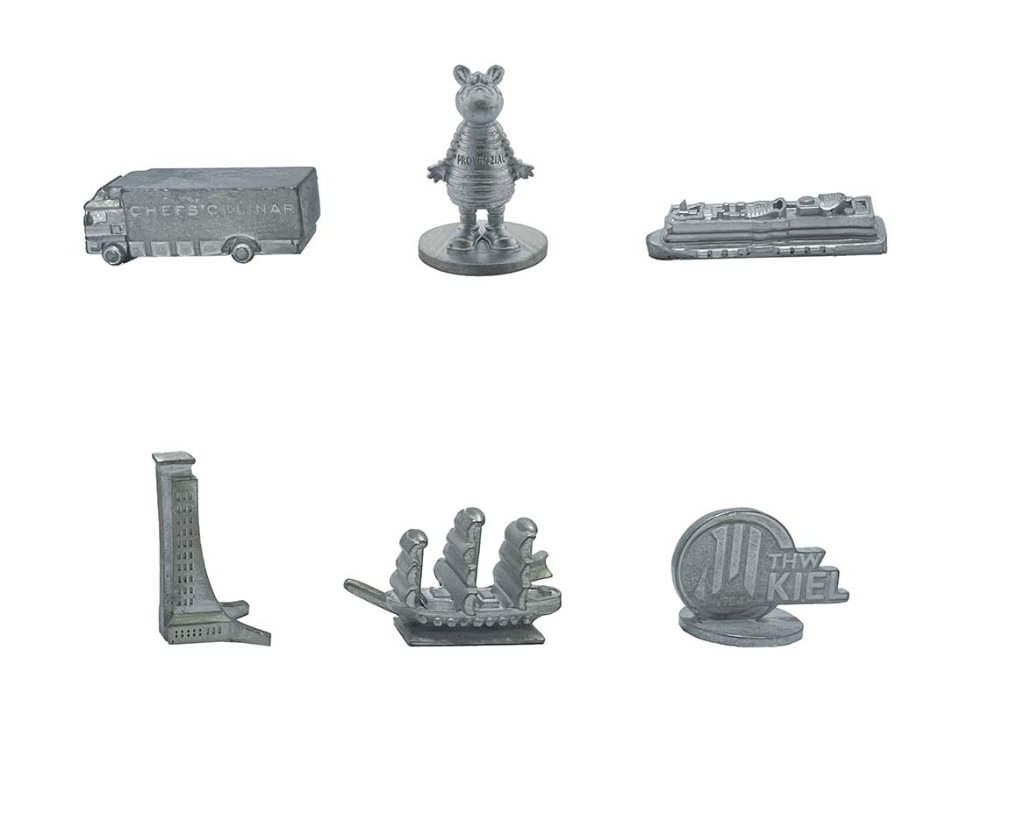 Sechs silberne Metallplättchen aus der Monopoly Kiel - City Edition sind abgebildet: ein LKW, ein Zebra, ein Kreuzfahrtschiff, ein Hochhaus, ein Segelschiff und ein THW Kiel Plättchen. Das Set ist auf einem weißen Hintergrund abgebildet.