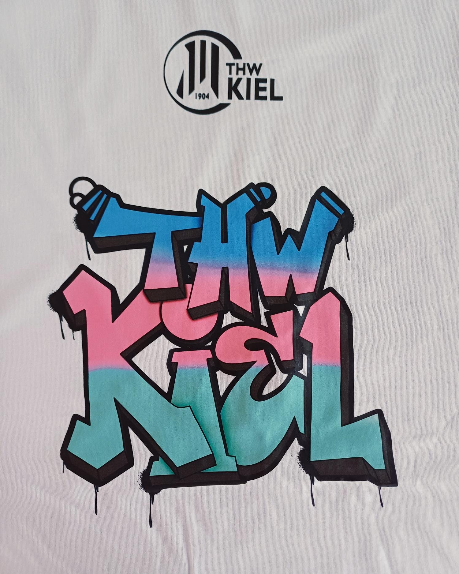 Das THW Kiel Tee Graffiti PUMA White besteht aus recycelter Baumwolle und zwei THW Kiel Prints: ein schwarzes, rundes Logo oben und auffällige Graffiti-Buchstaben in Pink, Blau und Schwarz mit Farbklecksen unten.