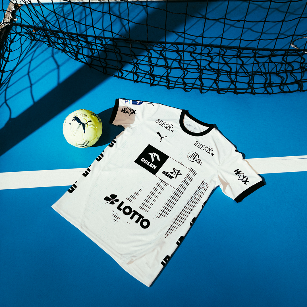Das THW Kiel 24-25 Heimtrikot PUMA Weiß mit schwarzen Zebra-Akzenten und Sponsorenlogos liegt auf einem blauen Spielfeld neben einem gelb-grünen PUMA-Fußball, während im Hintergrund Netzschatten erscheinen.