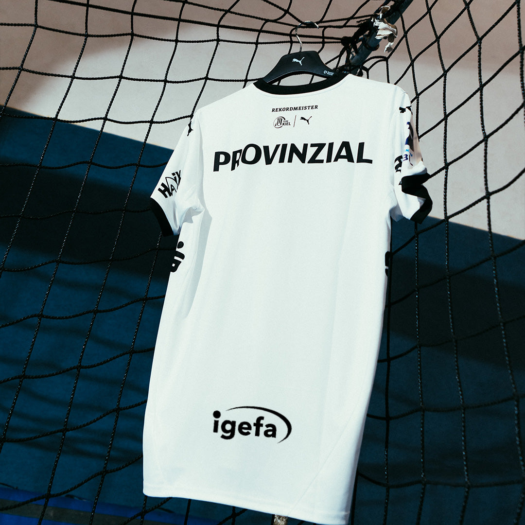 Das THW Kiel 24-25 Heimtrikot PUMA White zeigt ein klassisches Zebra-Design mit schwarzem Rand, Sponsorenlogos wie PROVINZIAL und igefa sowie PUMA-Branding, das vor einem lebendigen Sporthintergrund am Netz hängt.