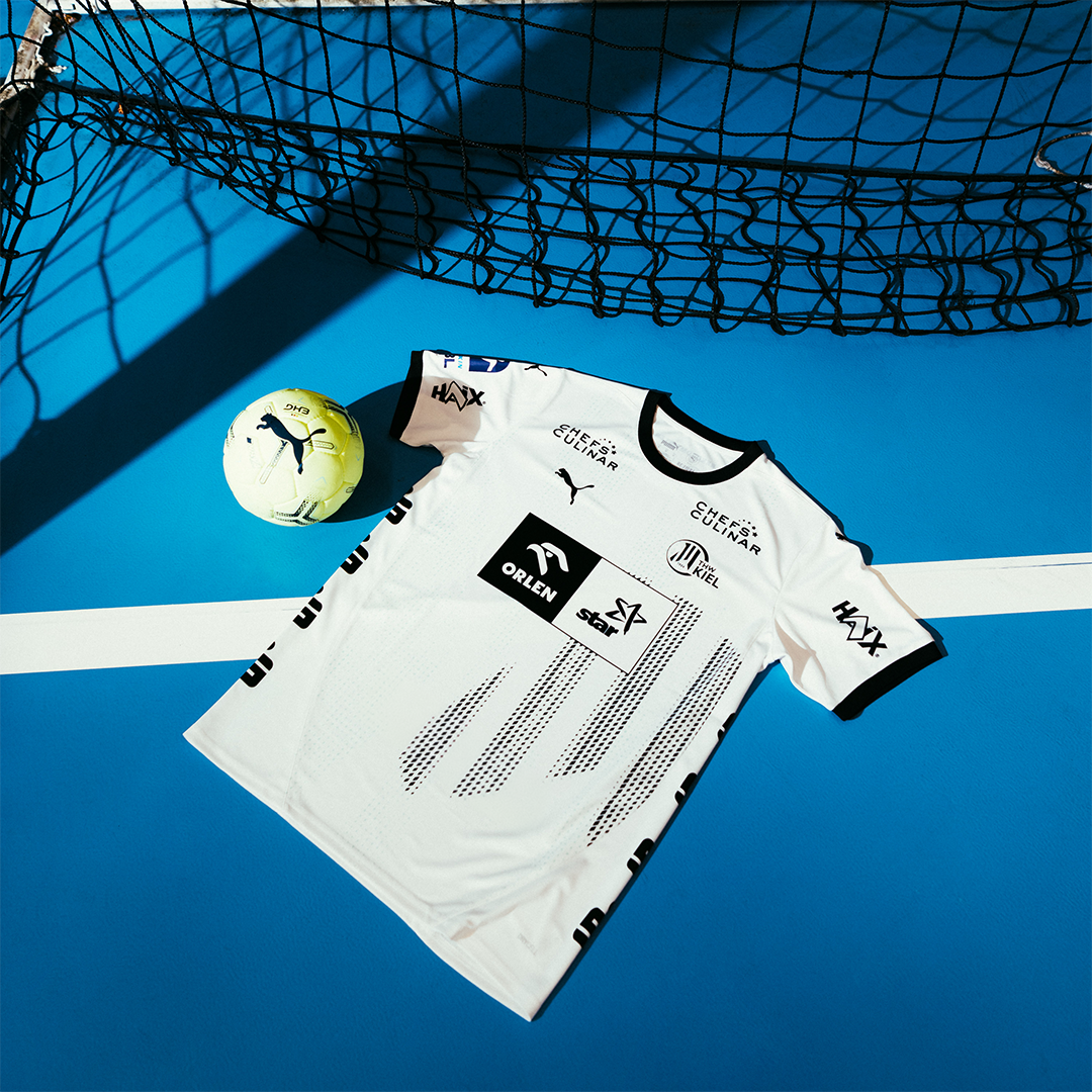 Ein THW Kiel 24-25 Heimtrikot PUMA Weiß mit Zebramuster liegt auf einem hellblauen Spielfeld neben einem gelben PUMA-Fußball, während Schatten von einem schwarzen Netz auf die lebendige Szene fallen.