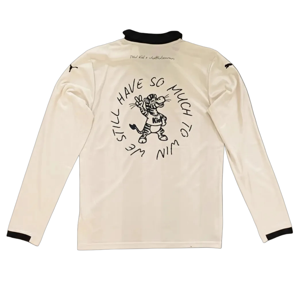 Das THW x whatthabeawears Jersey LS ist ein cremefarbenes Langarmshirt mit schwarzem Kragen und Manschetten, auf dem ein Cartoon-Tiger und der Text "We still have so much to win" zu sehen sind. Hergestellt aus recyceltem Polyester.