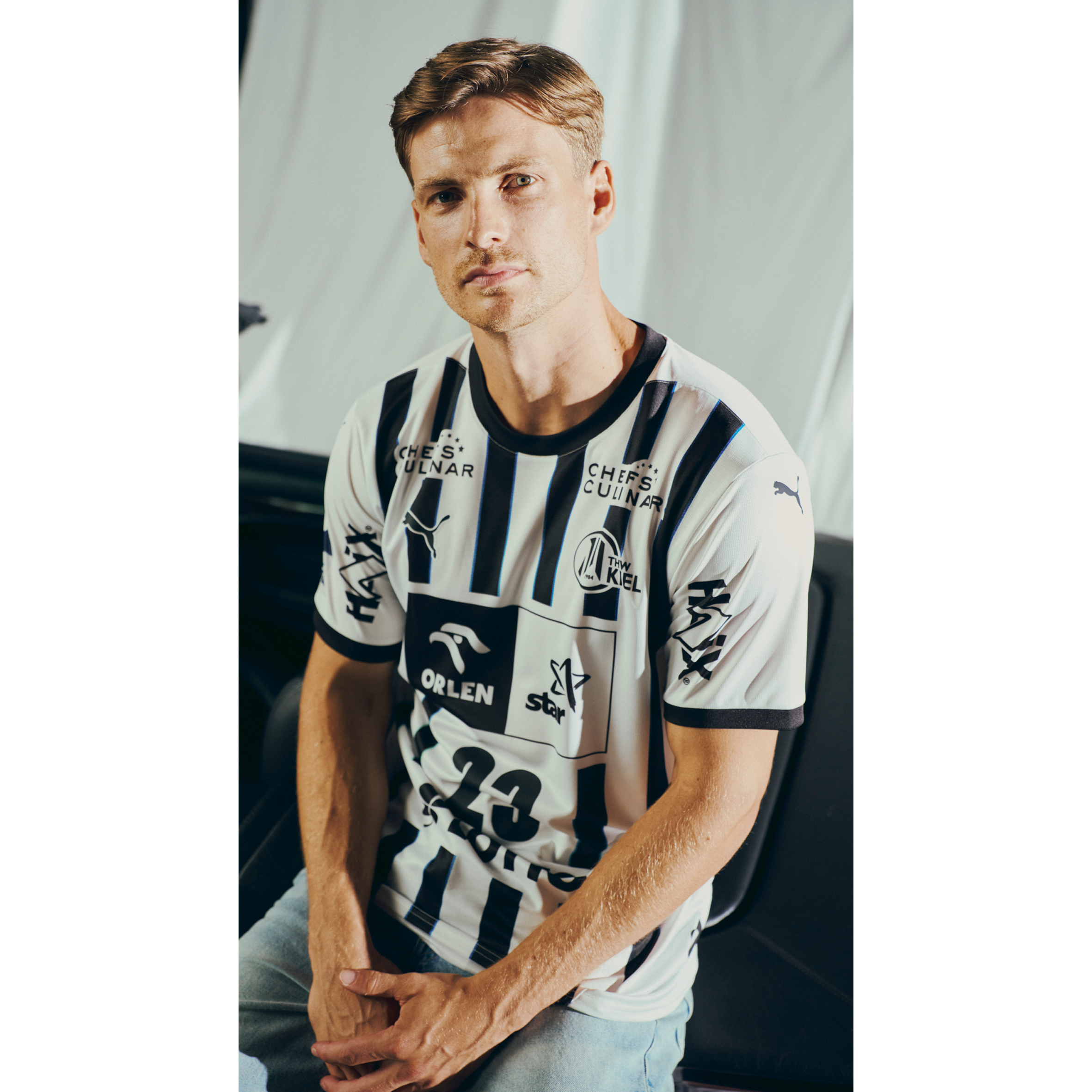 Ein Mann mit kurzen hellbraunen Haaren sitzt auf einer schwarzen Fläche, trägt das THW Kiel 25-26 Heimtrikot PUMA Weiß und hellblaue Jeans vor einem schlichten, hellen Hintergrund.