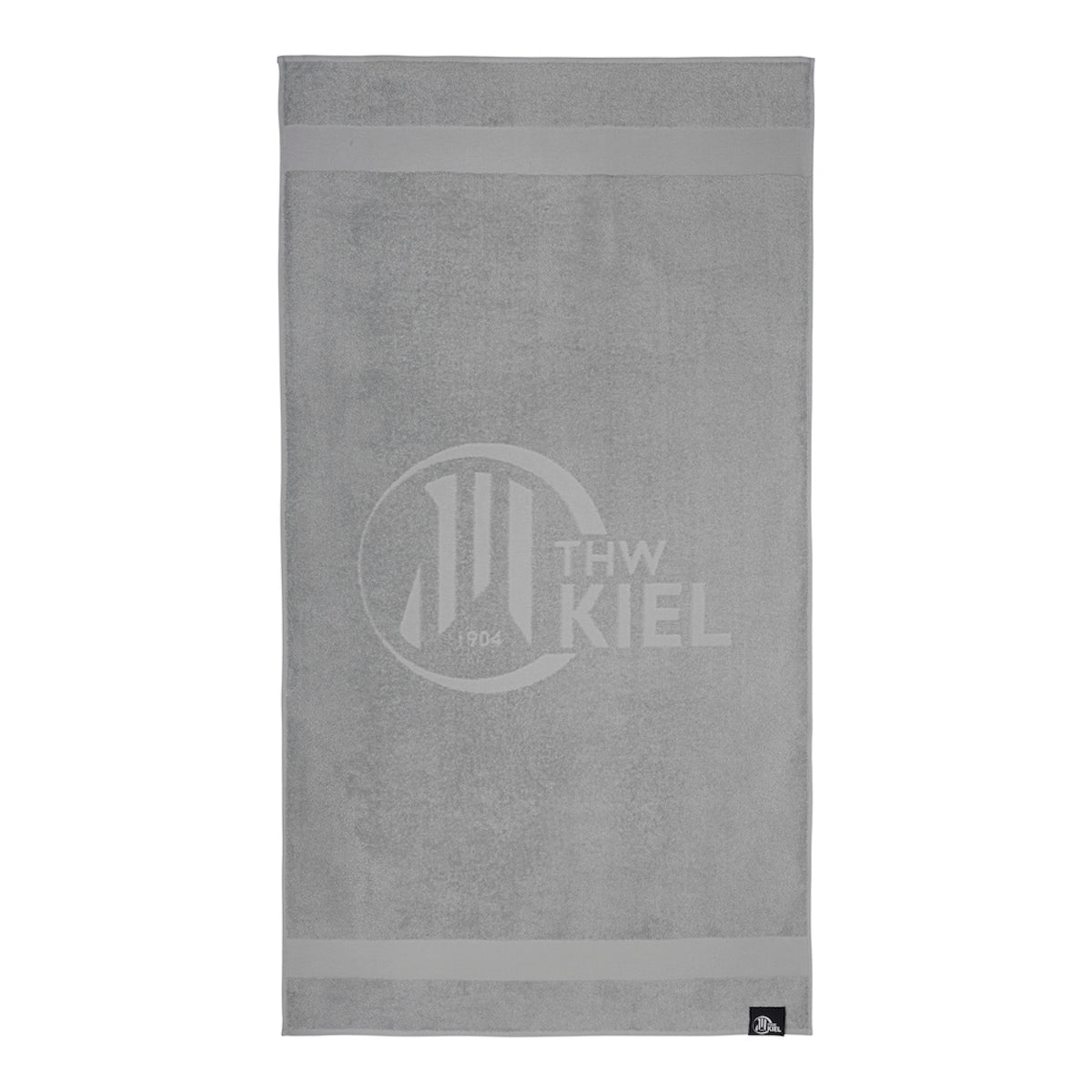 Das Badetuch hellgrau ist ein hellgraues, rechteckiges Handtuch mit dem Logo des THW Kiel und dem dezenten Aufdruck "Handball Rekordmeister 1904" in der Mitte, zwei horizontalen Streifen oben und unten und einem kleinen schwarzen Anhänger in der rechten unteren Ecke.