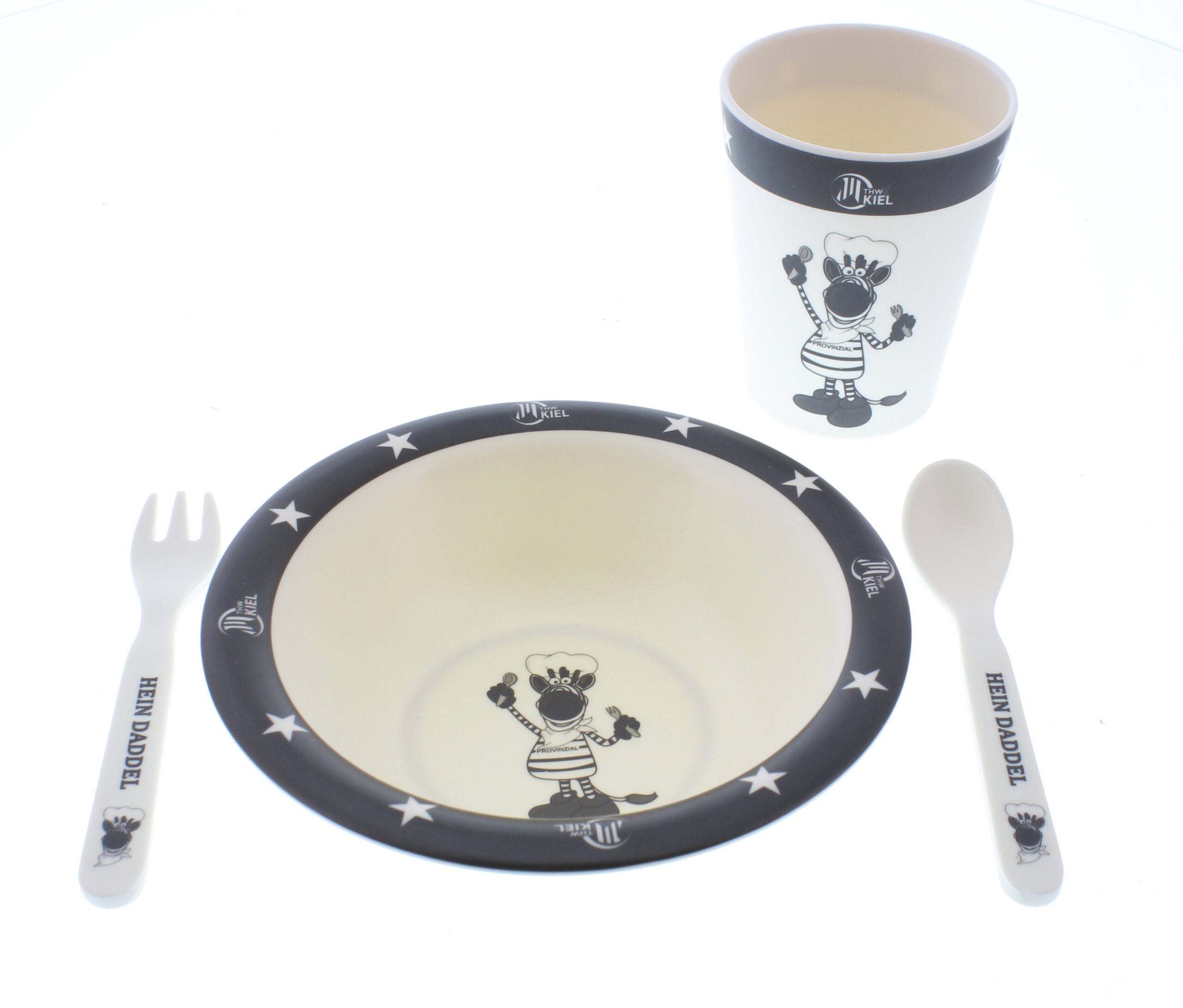 Das Geschirr-Set Kids 4-teilig weiß-schwarz ist ein Geschirr-Set für Kinder aus 100% Melamin mit Tasse, Schüssel, Löffel und Gabel mit Zebras, Sternen und dem Schriftzug "Hein Daddel" auf den Griffen.