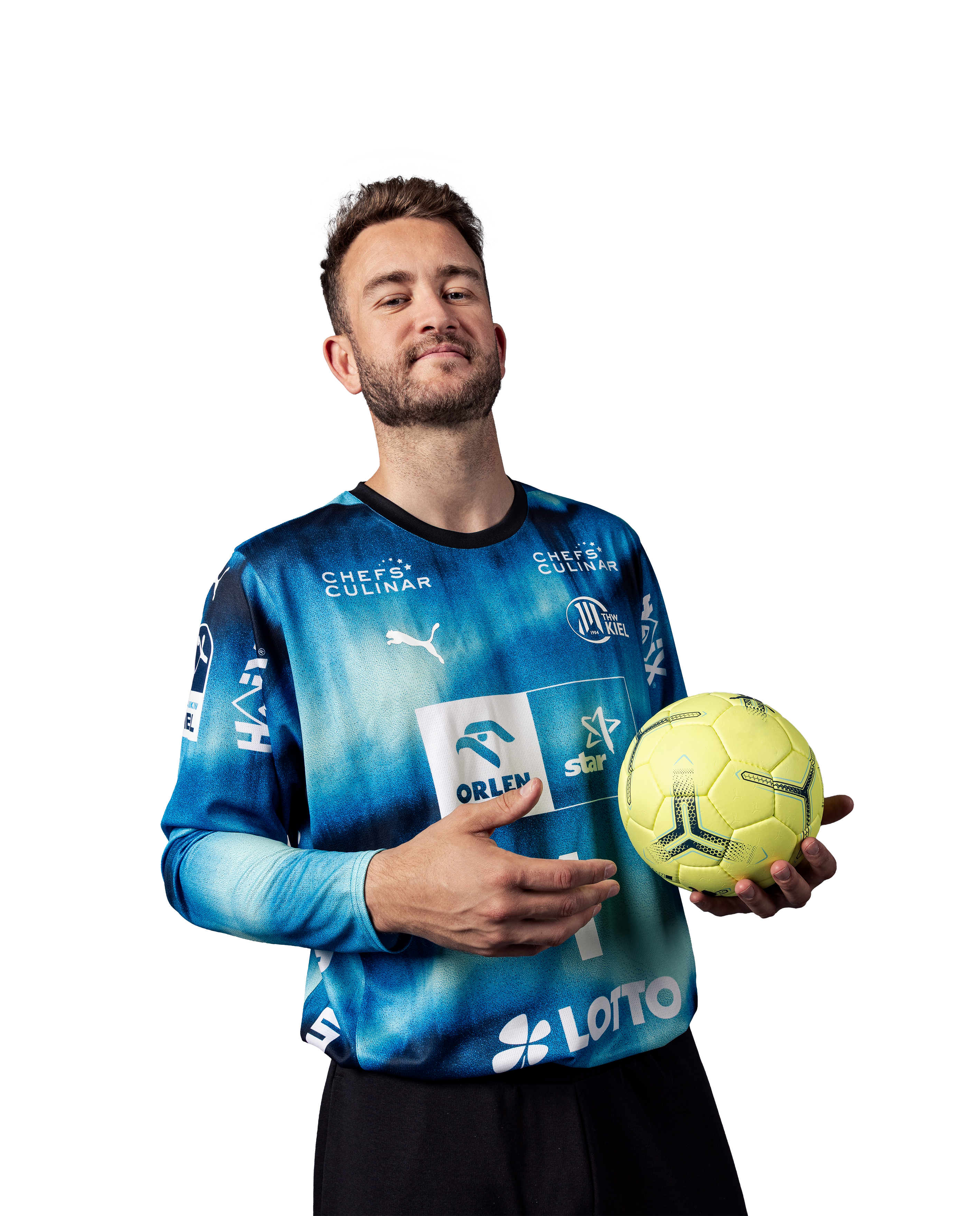 Ein Mann im THW Kiel 25-26 GK Trikot Home by PUMA in navy, mit Sponsorenlogos, hält einen gelben Fußball in der linken Hand. Er hat kurze Haare, einen leichten Bart und steht vor einem schlichten weißen Hintergrund.