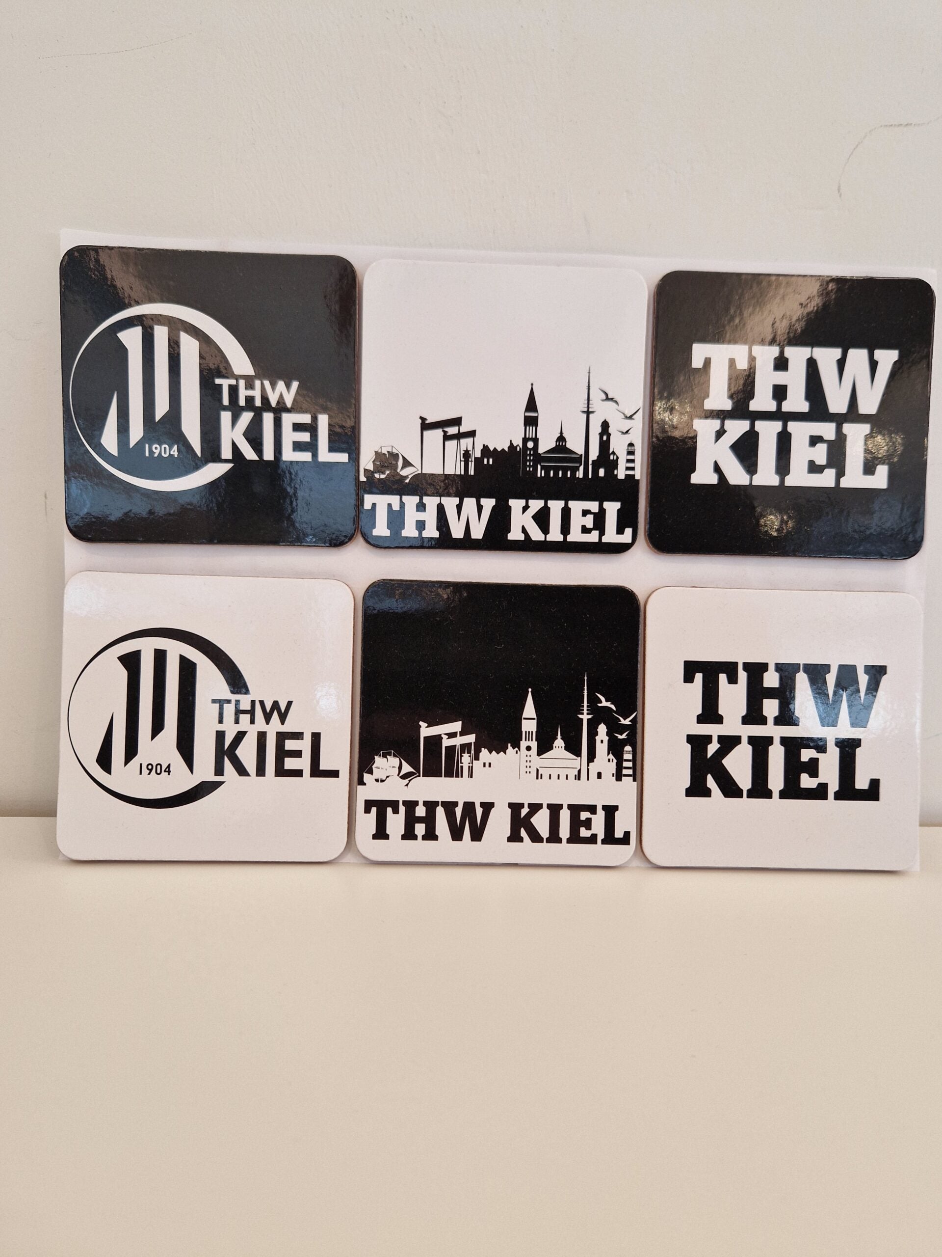 Das Set Untersetzer 6er schwarz-weiß enthält sechs rechteckige schwarz-weiße Untersetzer mit THW KIEL-Motiven, darunter das Logo, die Stadtsilhouette und der Schriftzug - ideal für die Sammlung eines jeden Handballfans.