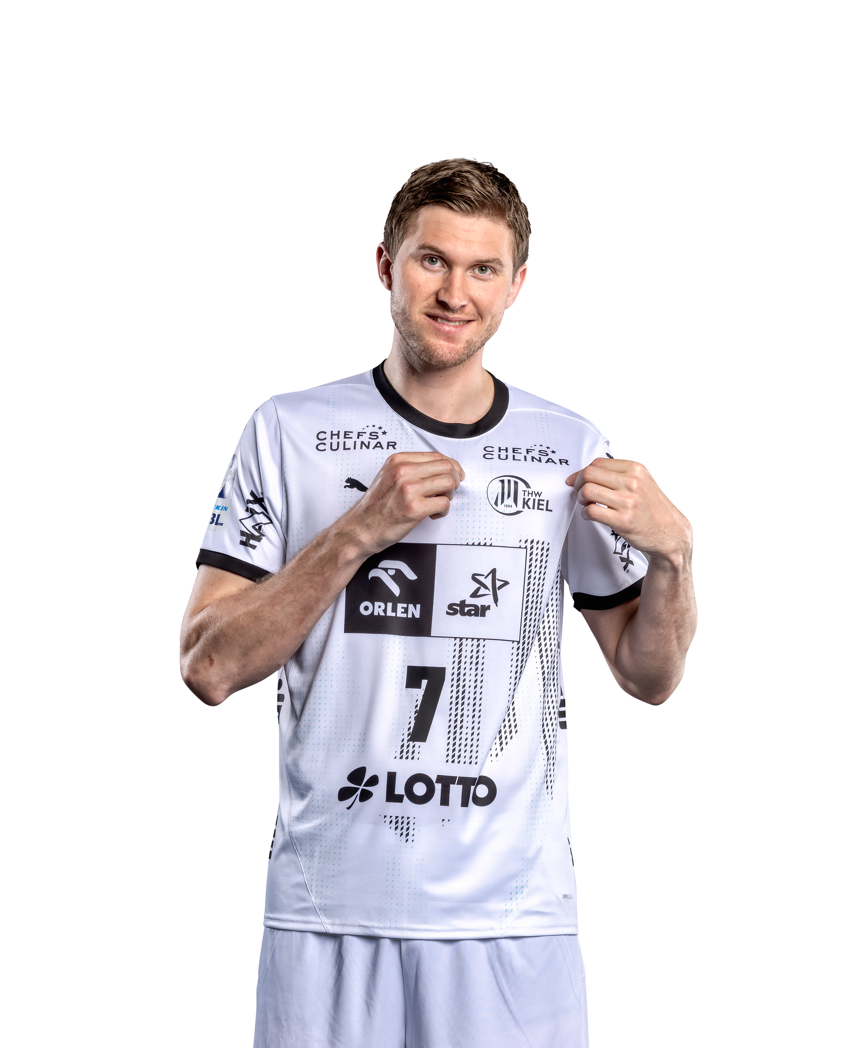 Ein männlicher Sportler, der das THW Kiel 24-25 Heimtrikot PUMA Weiß mit schwarzen Details und der Nummer 7 trägt, lächelt und zeigt auf das Teamlogo auf seiner Brust. Das Trikot zeigt mehrere Sponsorenlogos, die auf einem schlichten weißen Hintergrund angebracht sind.