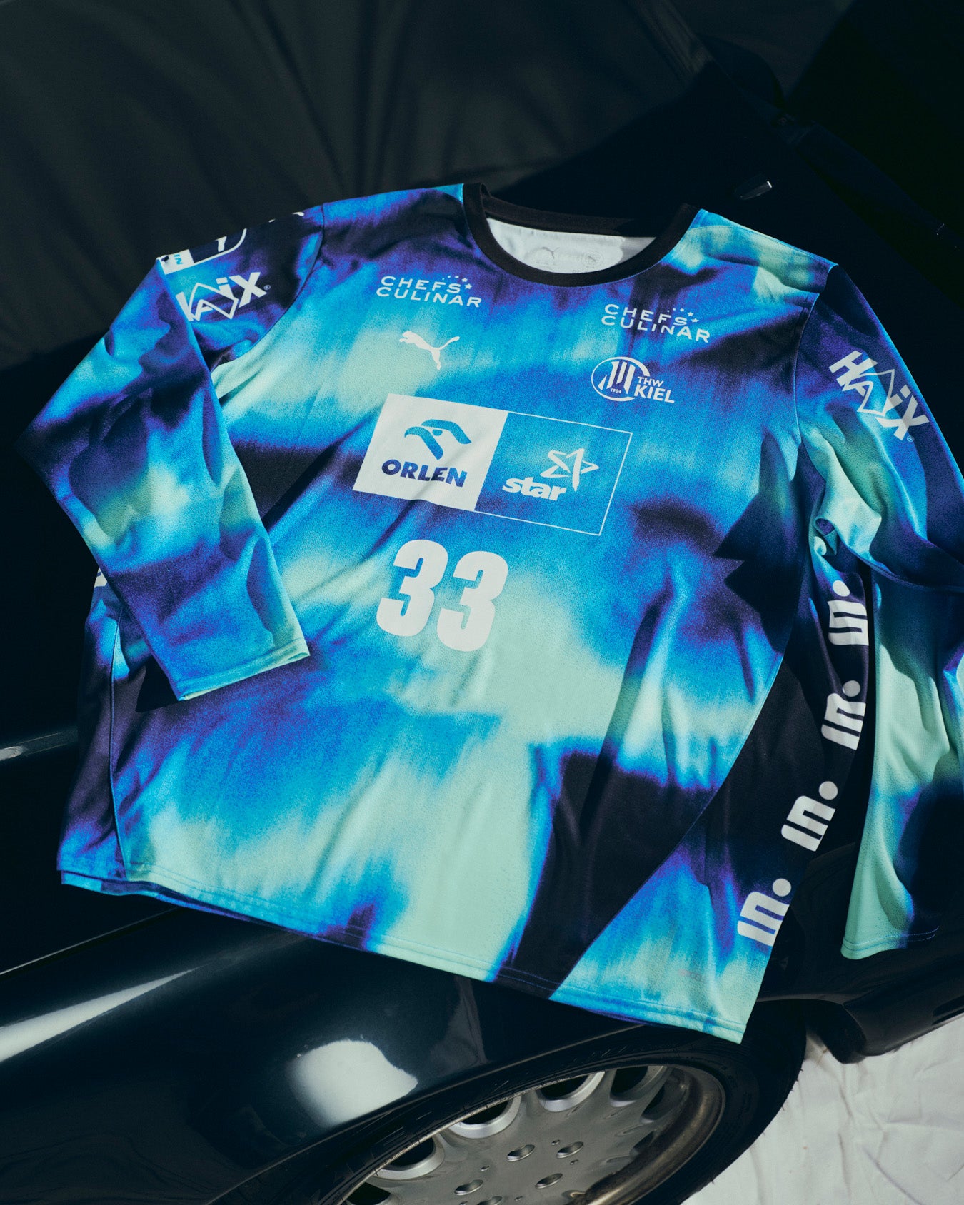 Das THW Kiel 25-26 GK Trikot Home Jr by PUMA in navy mit Sponsorenlogos und der Nummer 33 ist auf einer Motorhaube abgebildet, auf der das silberne Lenkrad zu sehen ist - und unterstreicht so den vom Nordlichter Design inspirierten Stil.