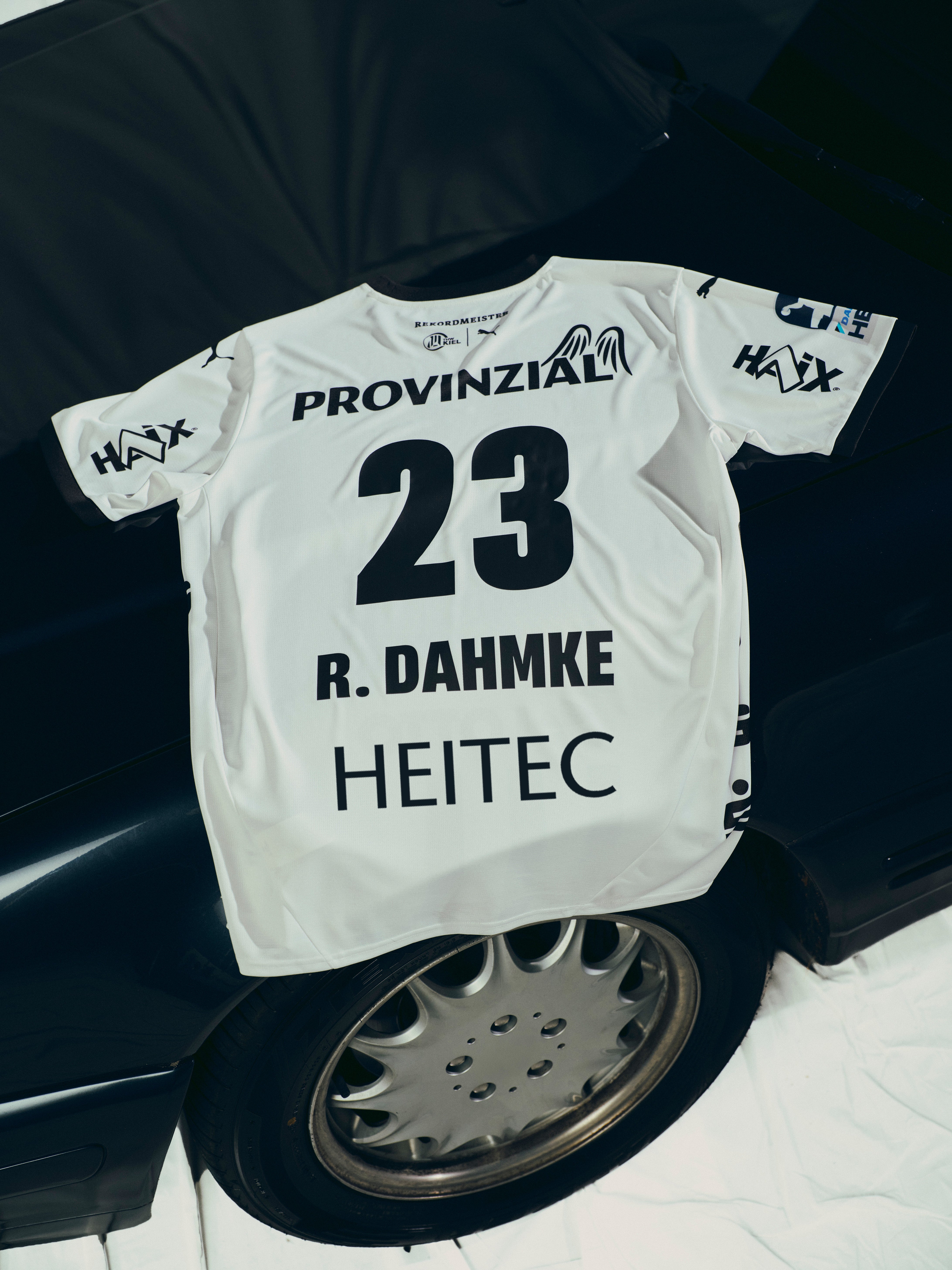Ein THW Kiel 25-26 Heimtrikot Jr PUMA White mit der Nummer 23 und R. Dahmke, das mit Sponsorenlogos wie PROVINZIA und HEITEC versehen ist, wird über ein schwarzes Auto drapiert - ein echtes Highlight für Zebras-Fans.