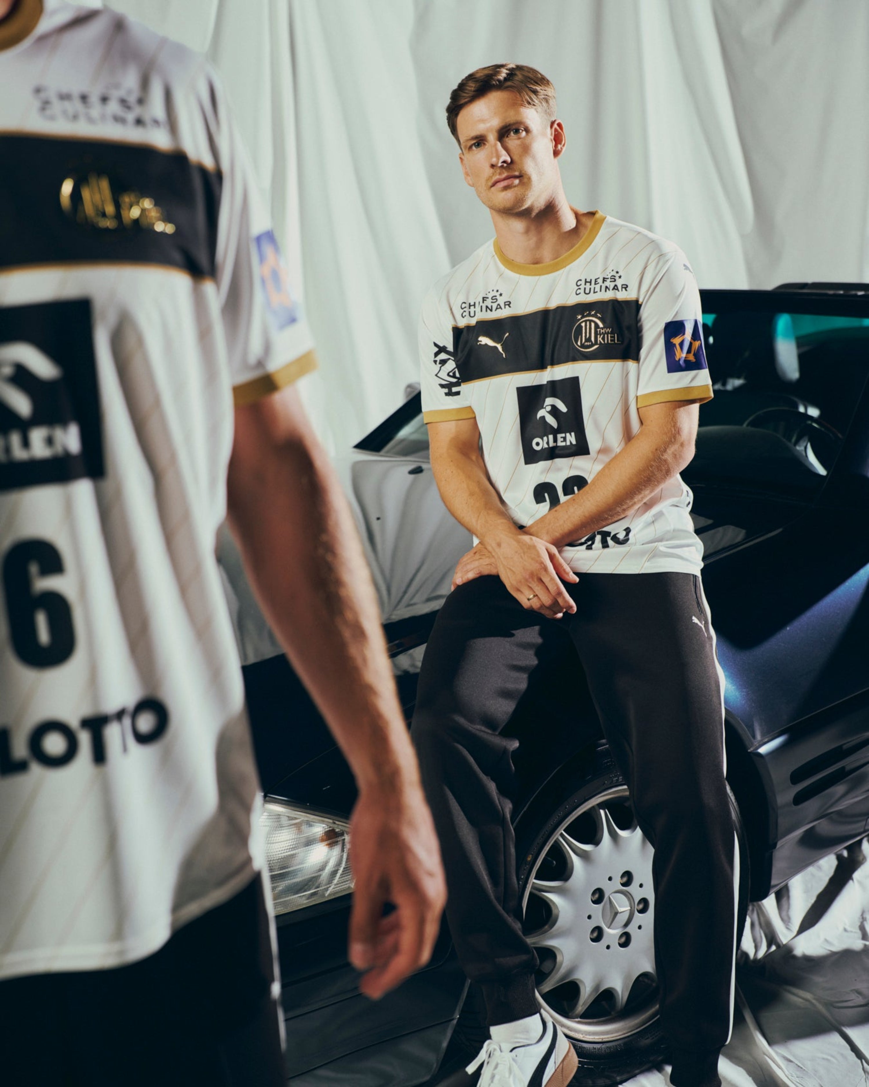 Ein Mann, der das THW Kiel 25-26 Euro Home Jersey Jr by PUMA in Weiß und Schwarz trägt, sitzt auf der Motorhaube eines dunklen Autos und schaut in die Kamera. Eine weitere Person in einem ähnlichen Trikot ist teilweise im Vordergrund zu sehen.