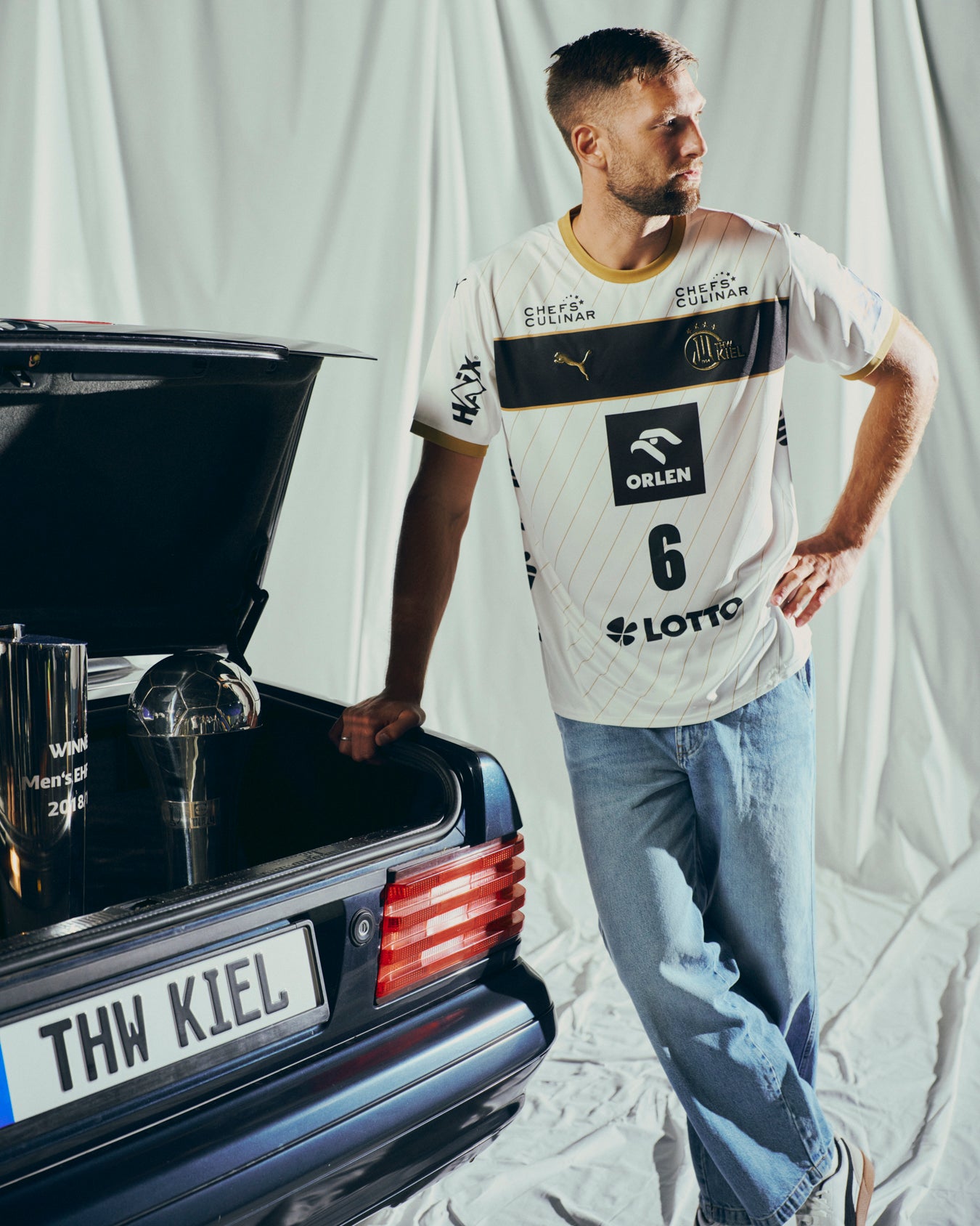 Ein Mann in einem weißen THW Kiel 25-26 Euro Home Jersey PUMA White und Jeans lehnt an einem schwarzen Auto mit THW KIEL-Kennzeichen. Der geöffnete Kofferraum offenbart einen Pokal vor einem Hintergrund aus weiß drapiertem Stoff.