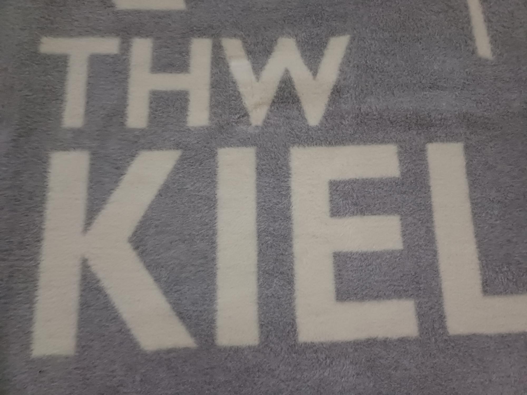 Eine grau-beige Kuscheldecke Linien mit fettgedrucktem, weißem THW KIEL Schriftzug quer darüber.