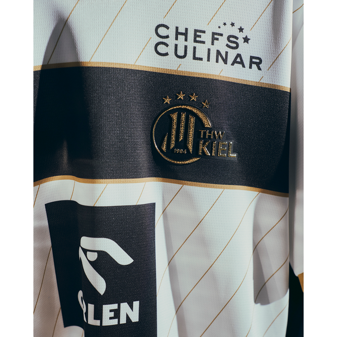 Eine Nahaufnahme des THW Kiel 25-26 Euro Home Trikots von PUMA in Weiß mit goldenen und schwarzen Streifen, Mannschafts- und PUMA-Logo, drei goldenen Sternen sowie CHEFS CULINAR- und PLEN-Sponsorenlogos - ideal für Fans der EHF European League.