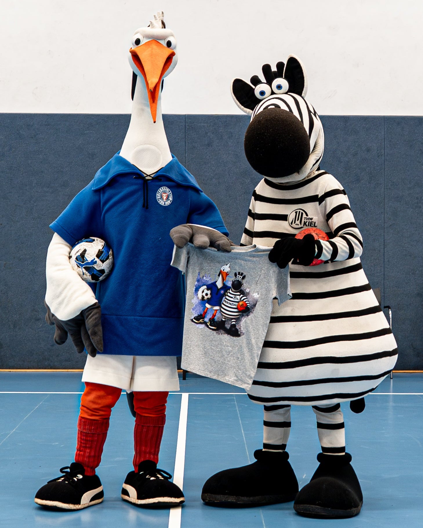 Zwei Maskottchen, das gestreifte THW-Zebra und der Holstein-Storch in einem blauen Trikot mit einem Fußball, stehen auf einem Hallenplatz. Das Zebra trägt das T-Shirt „Kids Holstein Kiel x THW Kiel“, auf dem beide Maskottchen als Comicfiguren abgebildet sind.