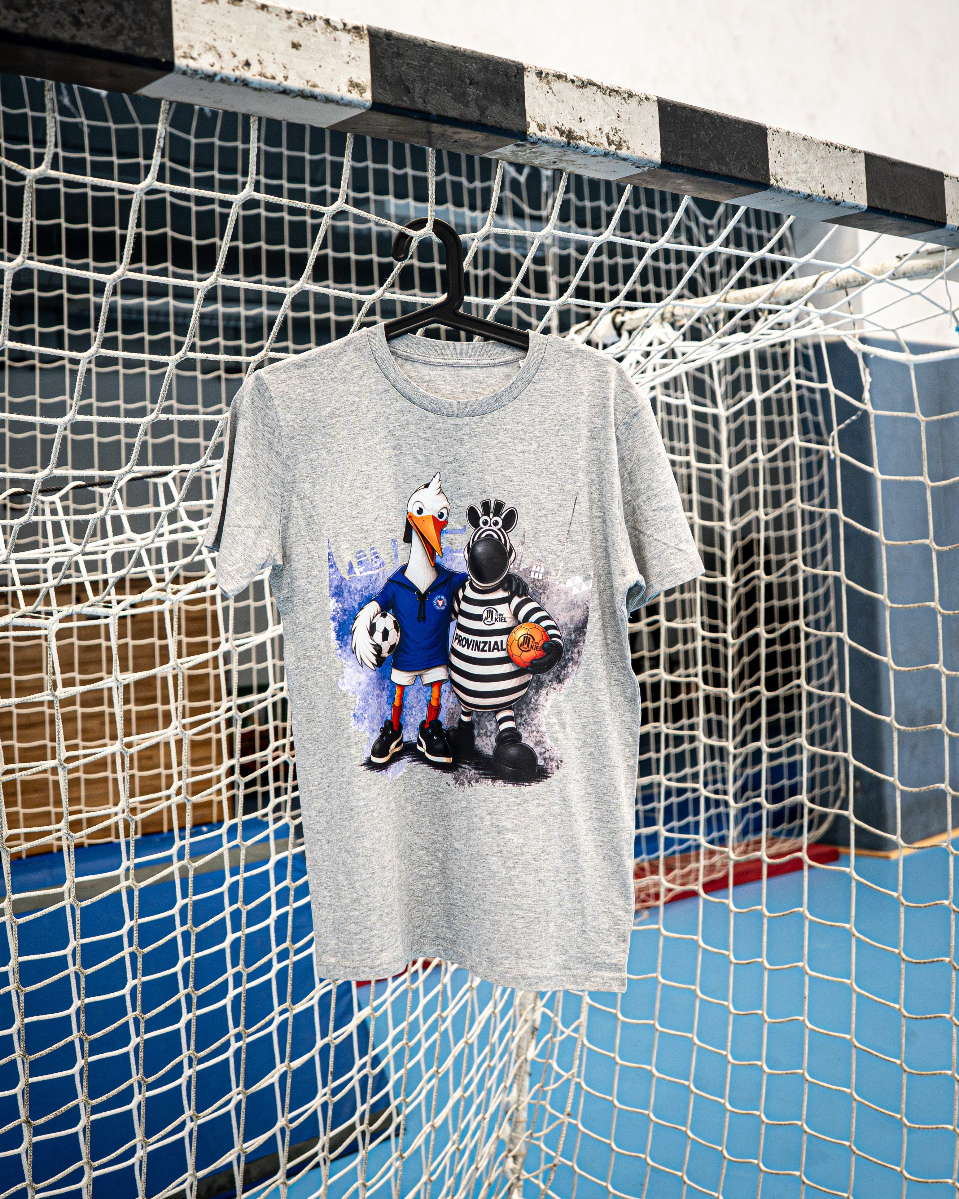 Ein graues Kinder-T-Shirt mit dem Aufdruck „Holstein Kiel x THW Kiel“ und den Comic-Maskottchen – einem Storch mit einem Fußball und einem Stier mit einem Basketball – hängt an einem Kleiderbügel am Tornetz in einer Sporthalle.