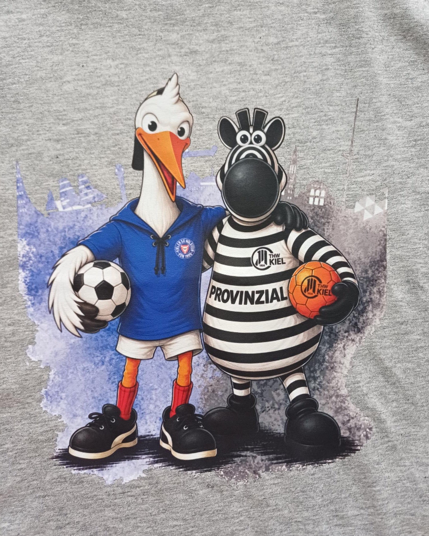 Ein fröhlicher Holstein-Storch in einem blauen Trikot mit einem Fußball steht neben dem schwarz-weißen THW-Zebra, das einen Handball in der Hand hält. Beide Maskottchen posieren gemeinsam – perfekt für das T-Shirt „Kids Holstein Kiel x THW Kiel“.