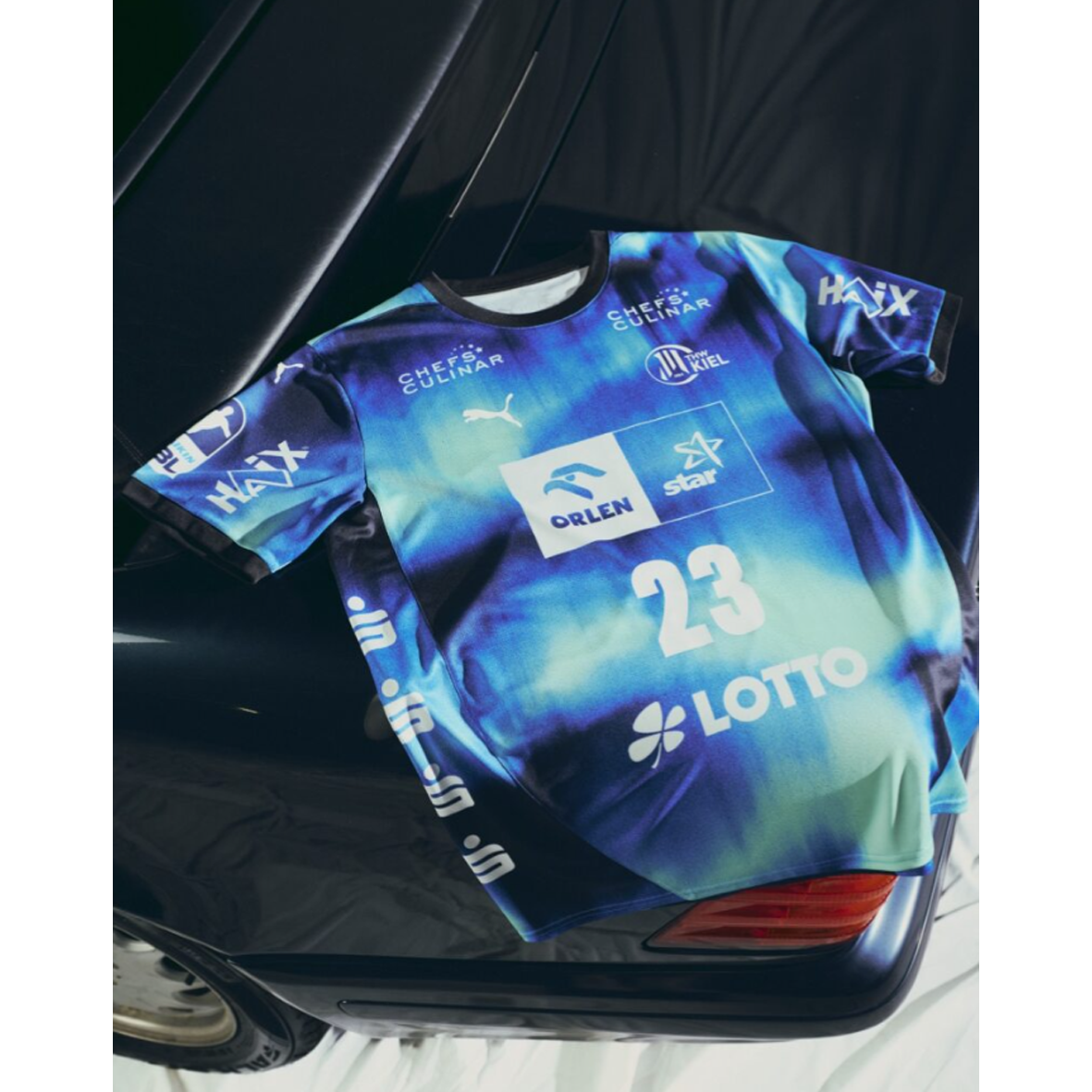 Das navyfarbene THW Kiel 25-26 Auswärtstrikot von PUMA mit Sponsorenlogos und der Nummer 23 ist über ein schwarzes Auto drapiert - ein echter Hingucker unter den modernen Trikots.