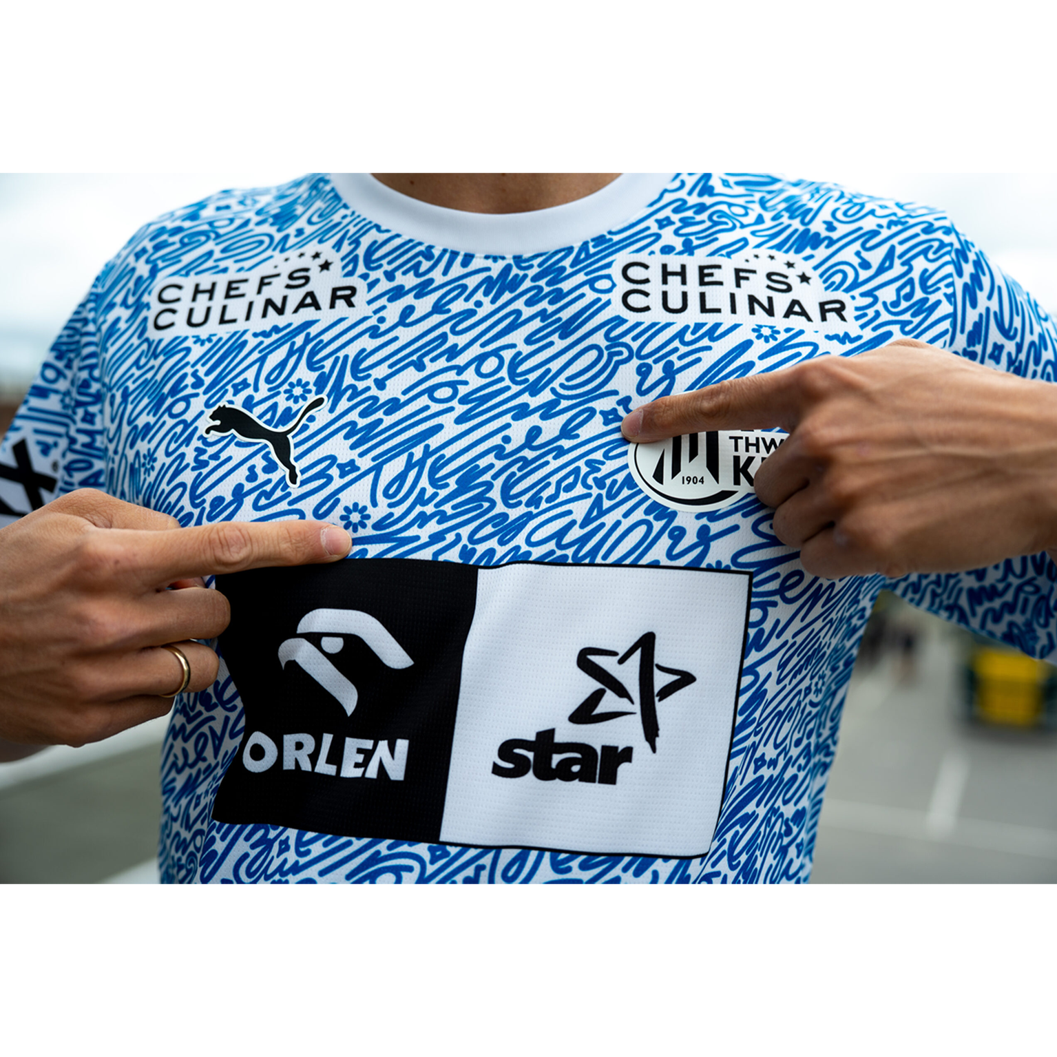 Eine Person, die das THW Kiel 24-25 Sondertrikot "Kieler Woche" Racing Blue, entworfen von Cihan Tamti mit abstrakten Mustern, trägt, zeigt auf das Wappen. Auf dem Trikot sind die Logos von Chefs Culinar, Puma, Orlen und Star abgebildet.