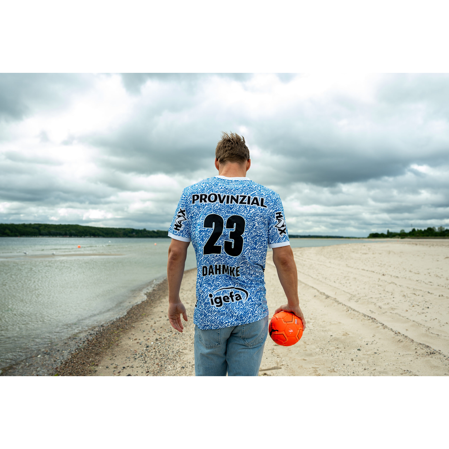 Eine Person in Jeans und einem blauen THW Kiel 24-25 Spezialtrikot "Kieler Woche" Racing Blue mit der Nummer 23 läuft über einen Sandstrand, in der Hand einen orangefarbenen Ball, mit Wasser und bewölktem Himmel im Hintergrund.