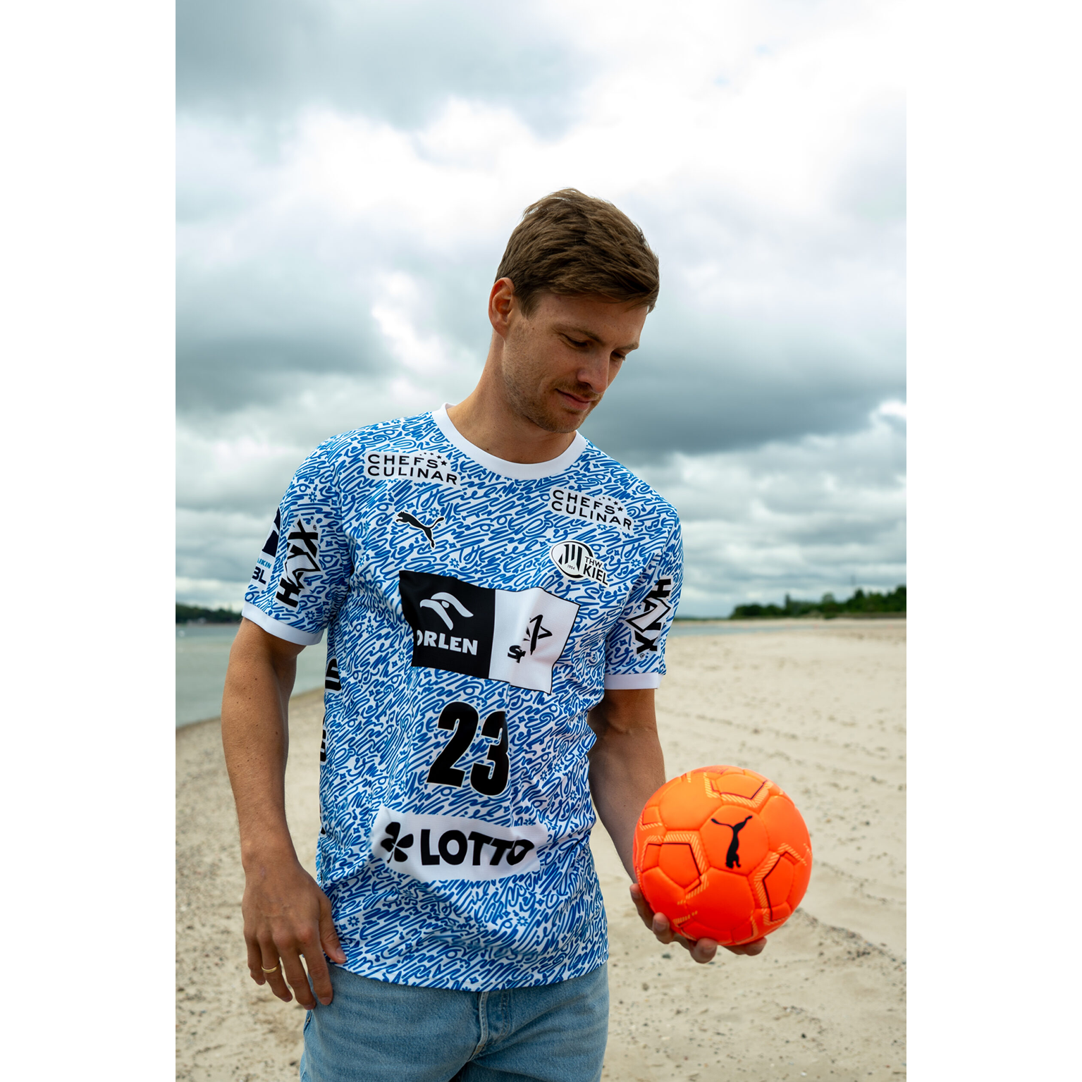 Ein Mann an einem Sandstrand unter einem bewölkten Himmel blickt nach unten, er hält einen orangefarbenen Fußball und trägt das THW Kiel 24-25 Special Jersey "Kieler Woche" Racing Blue mit Sponsorenlogos und der Nummer 23.