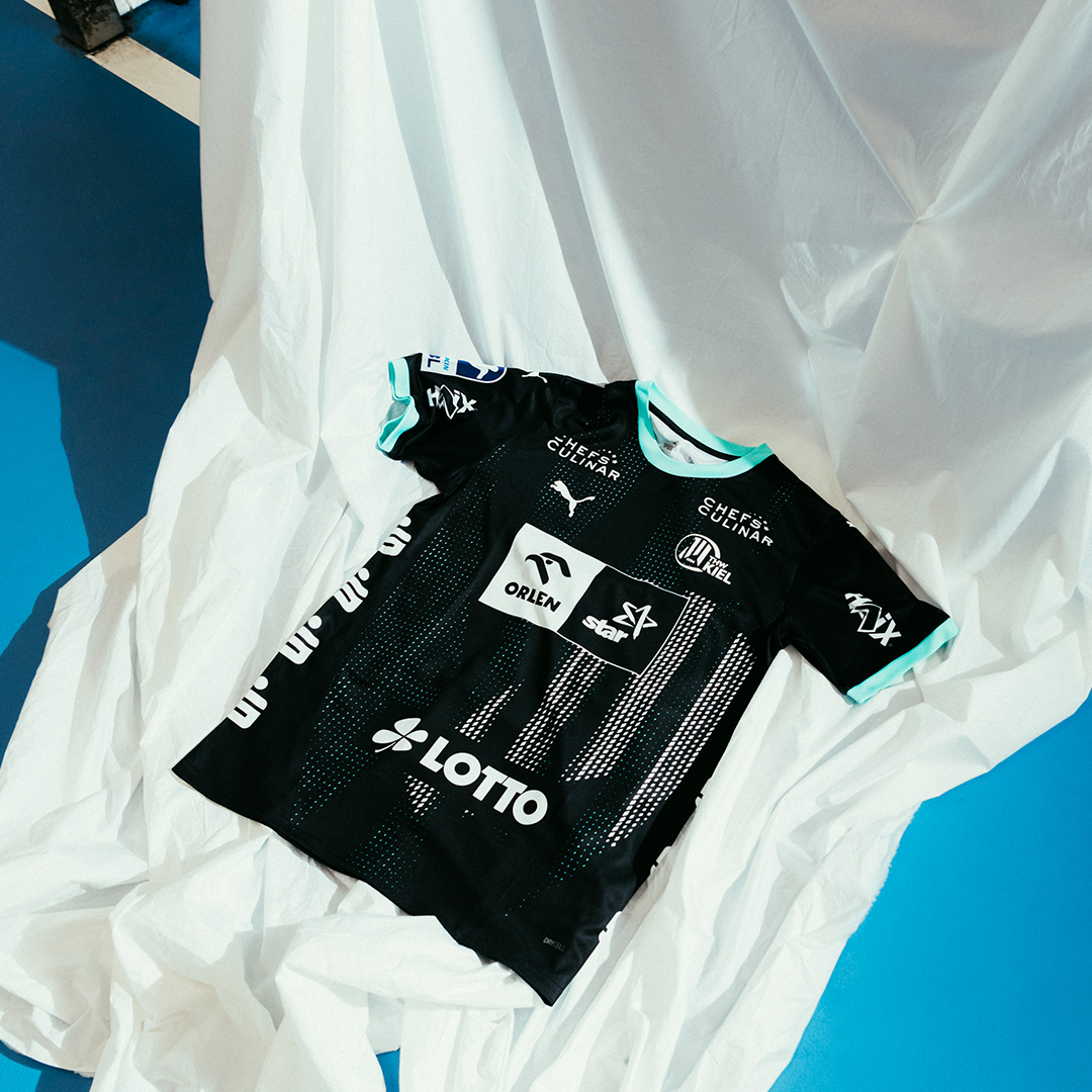 Das THW Kiel 24-25 Auswärtstrikot in PUMA-Schwarz mit tealfarbenem Rand und weißen Logos von Puma, Lotto und Orlen ist auf einem zerknitterten weißen Laken abgebildet, wobei der blaue Boden im Hintergrund teilweise sichtbar ist.